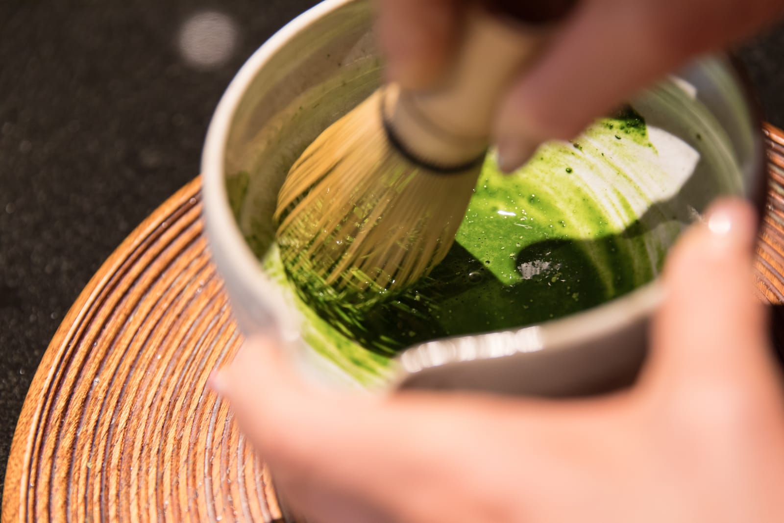 Close view of hands whisking bright green matcha with a bamboo whisk in a pale tea bowl, the bowl set on a turned wooden tray