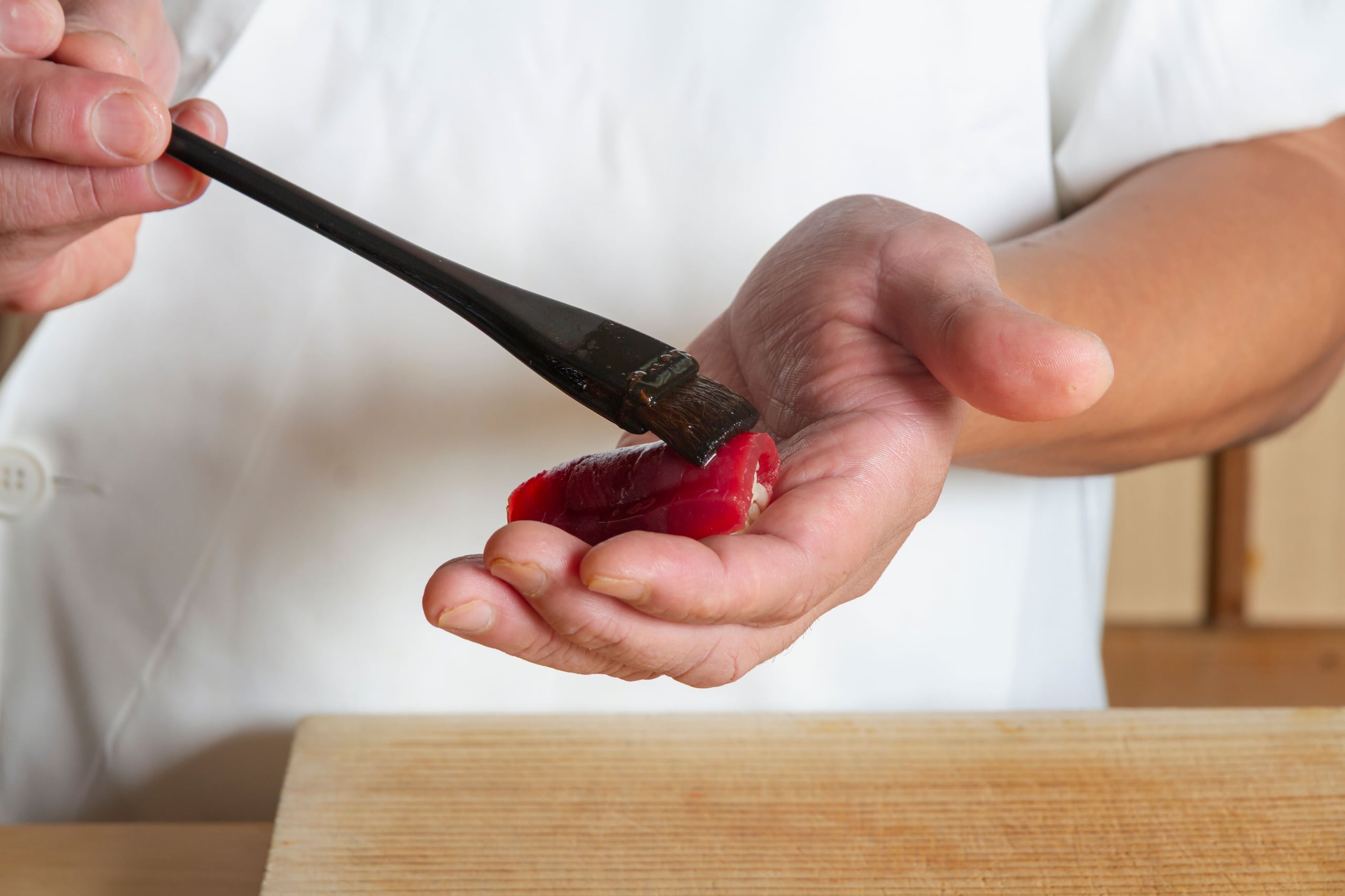 A sushi chef in a white uniform brushing nikiri soy glaze onto a piece of tuna held in the palm of his hand