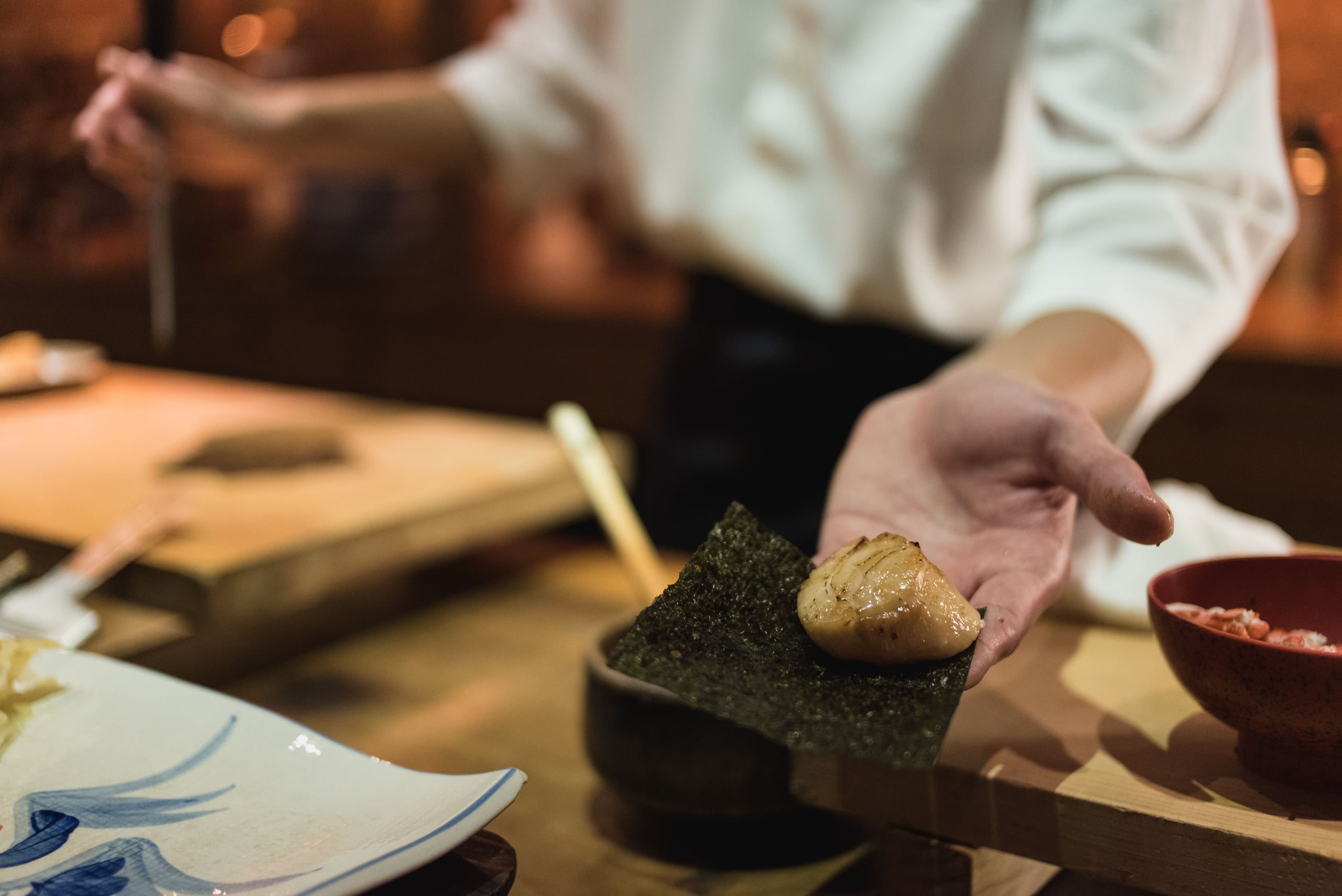 A chef across the counter presenting a seared scallop wrapped in a strip of nori