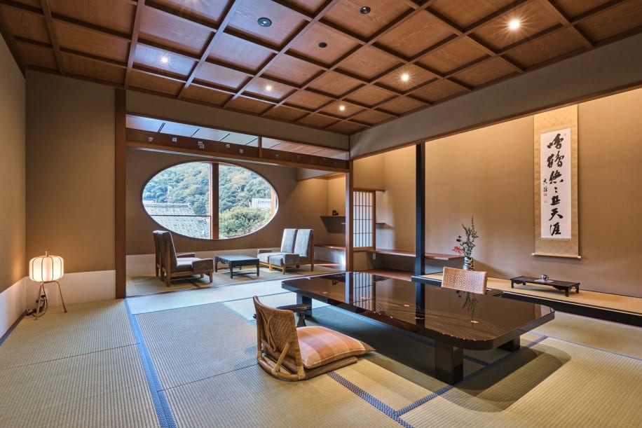 A tatami guest room in a Kyoto ryokan with a low window seat and a paper floor lantern, shoji screens open to a maple garden outside