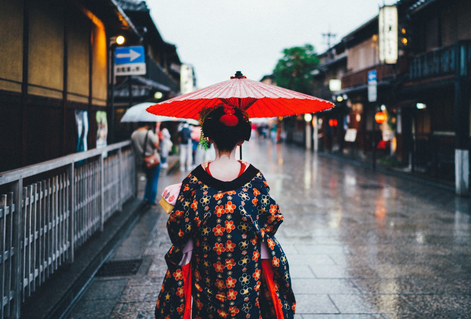 A figure in kimono carrying a red paper parasol walks away down a rain-wet Gion street lined with wooden facades and lanterns