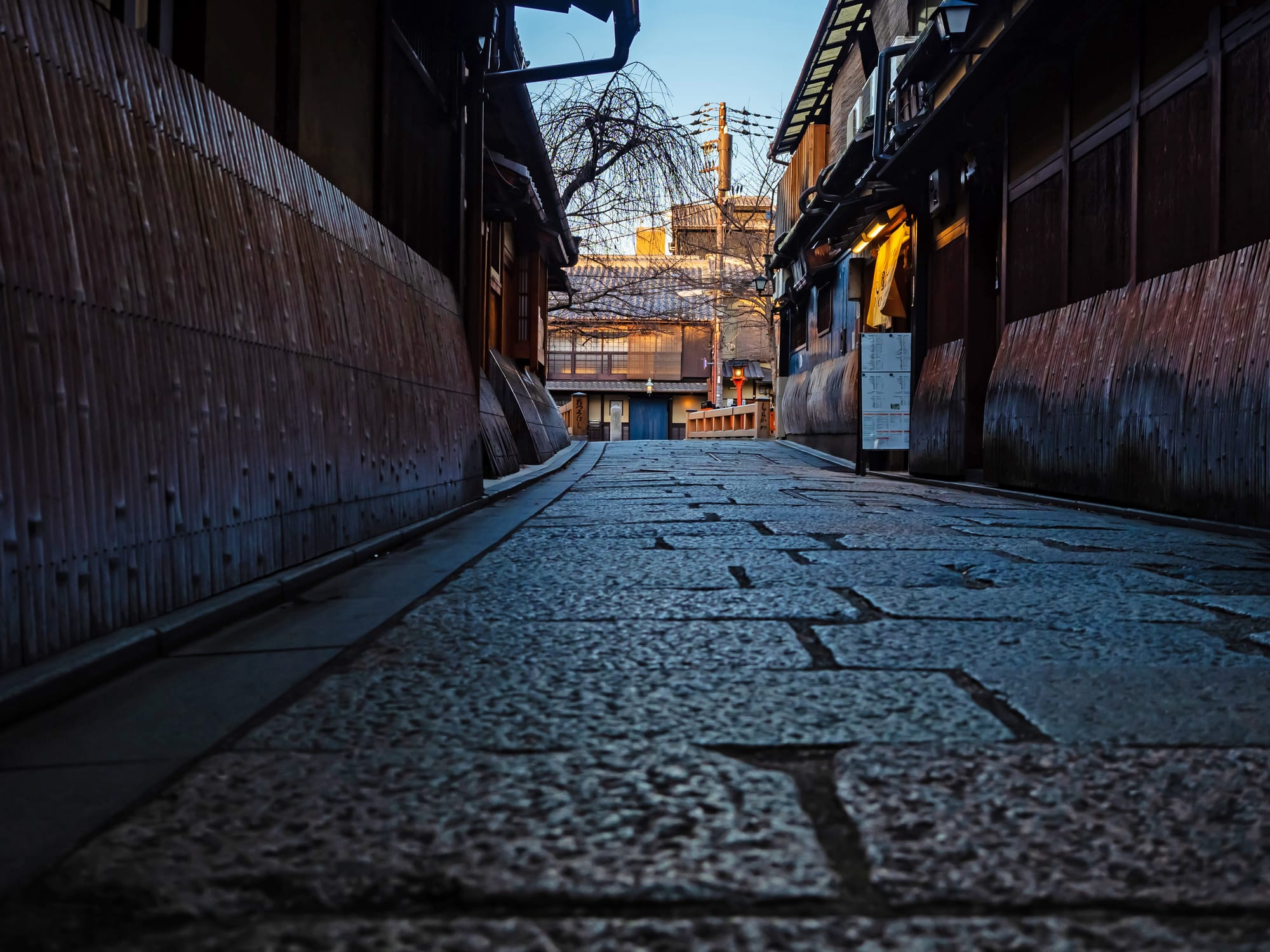 An empty cobblestone lane at dusk in Kyoto's Gion district, lined with dark wooden machiya facades and a single lit lantern