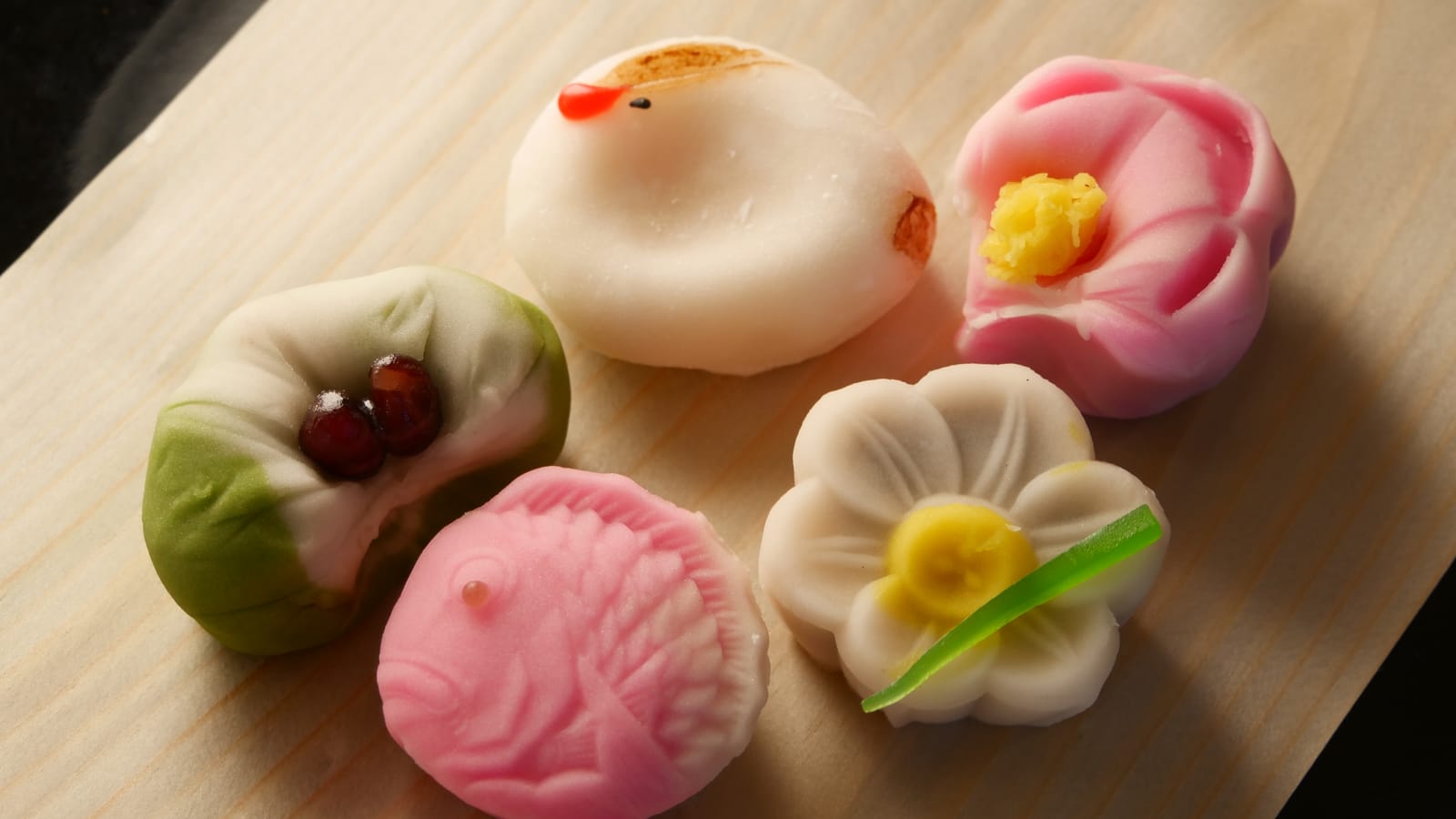 Five wagashi sweets on pale wood: a white bird, a pink camellia, a pink sea bream, a white narcissus and a green sweet filled with red beans