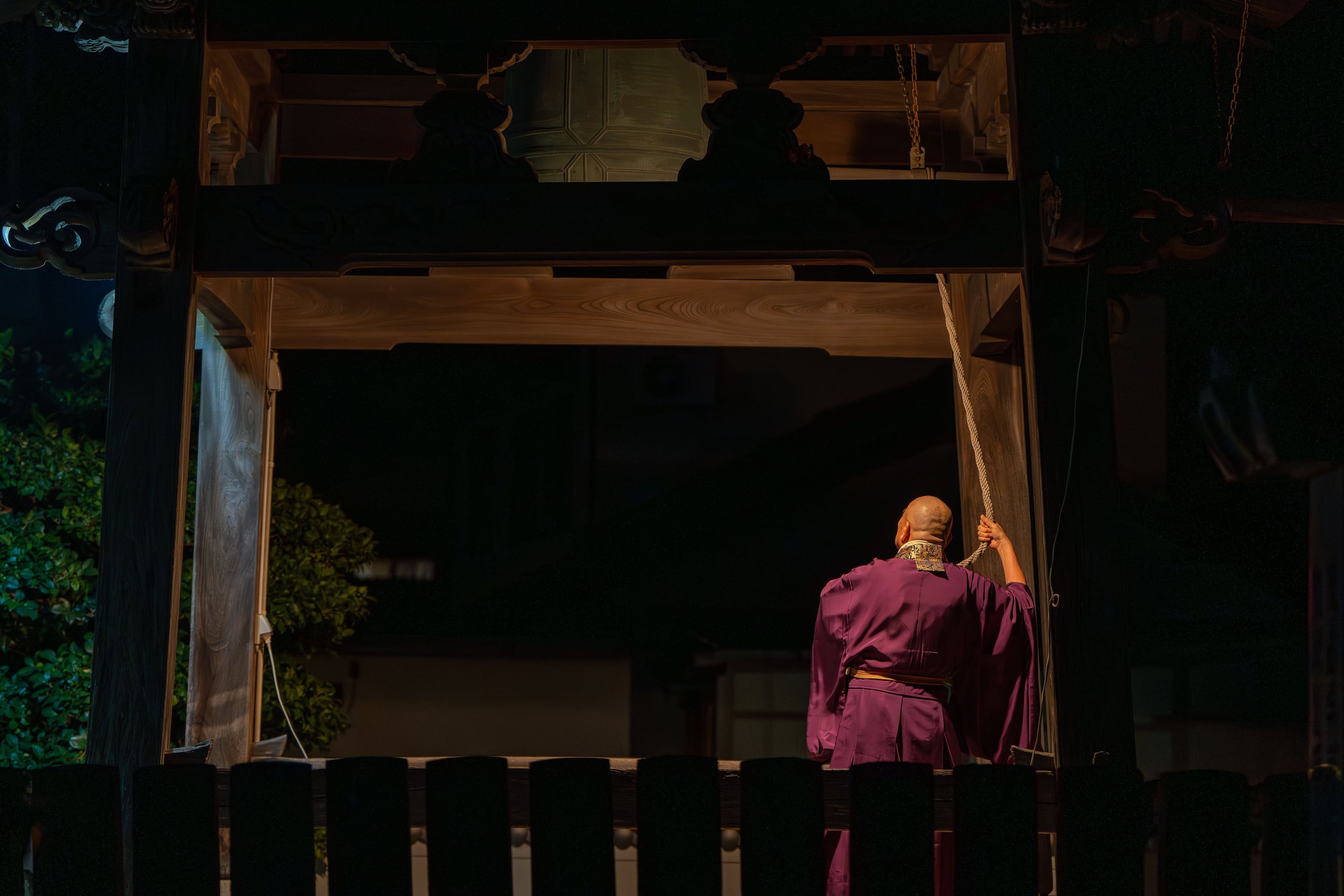 A monk in magenta ceremonial robes seen from behind, ringing a great temple bell