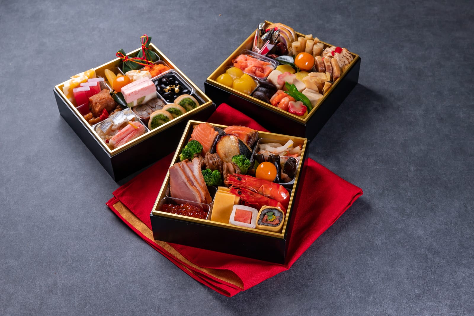 Three stacked lacquered jūbako boxes, each divided into compartments filled with osechi ryōri: shrimp, black beans, rolled kelp and other New Year dishes