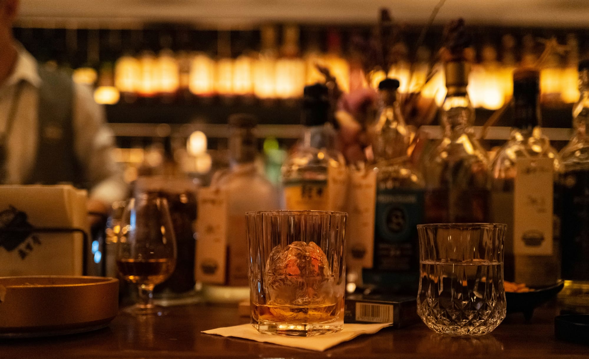 A tumbler of whisky over a single sphere of ice on a dark bar counter, a cut-glass tumbler of water beside it and bottles lit behind