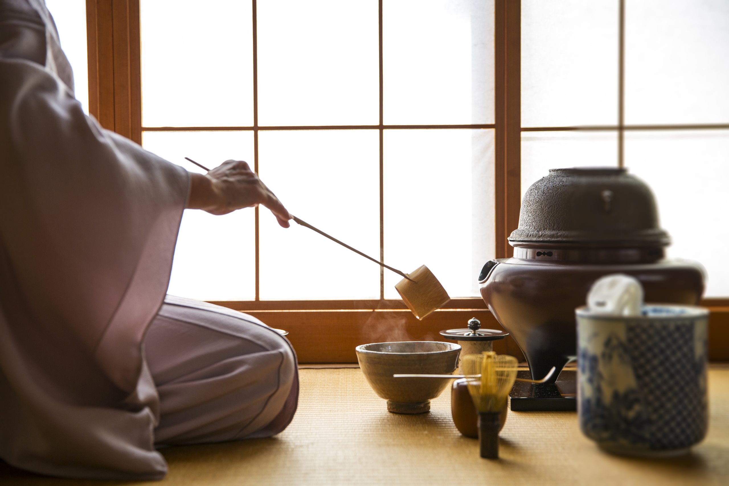 A host in a pale kimono kneels by a low window, ladling water for tea