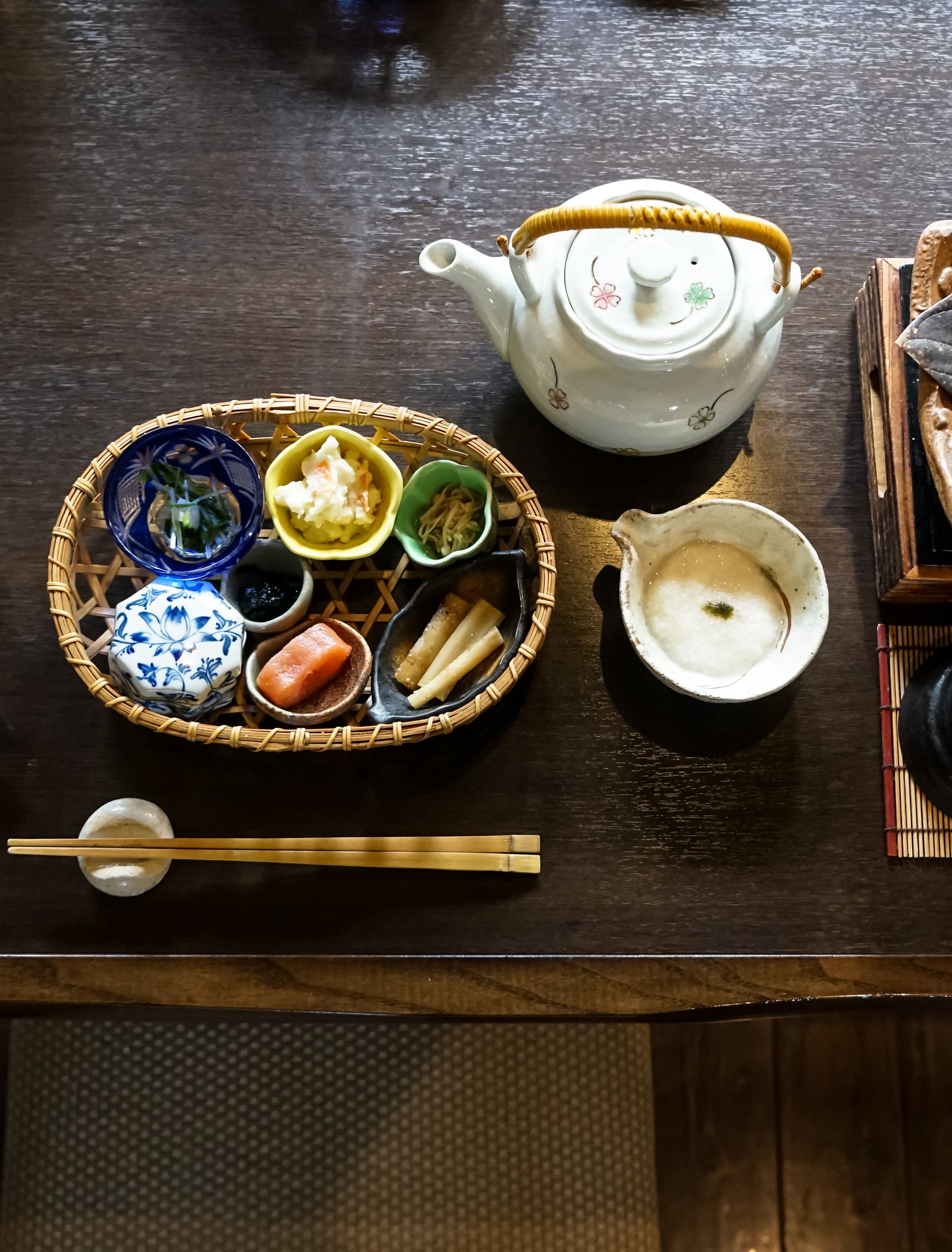 A Japanese breakfast laid on a dark wooden table: small dishes in a bamboo basket, a teapot and a cup beside it