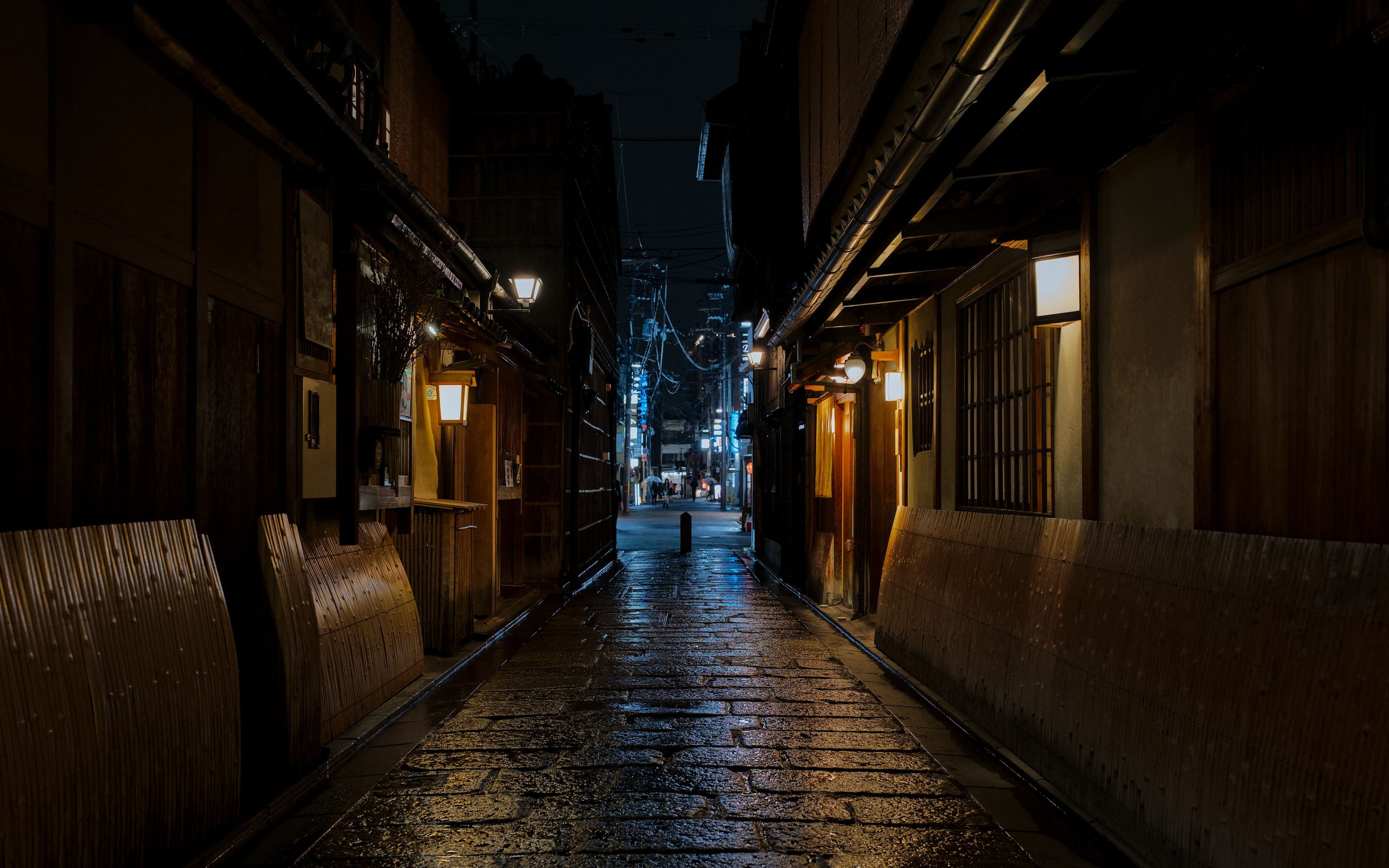 A narrow stone-paved lane in Gion at night after rain, lanterns lit along wooden teahouse fronts and bamboo fences
