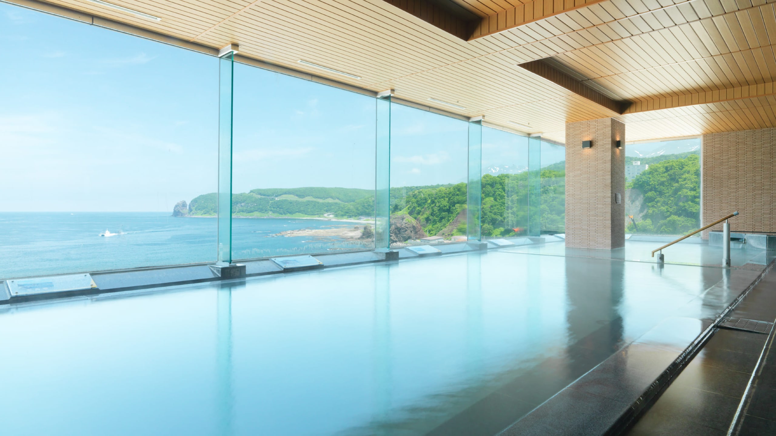 The long indoor onsen of Kitakobushi Shiretoko behind a wall of glass, the Okhotsk Sea and a green headland beyond