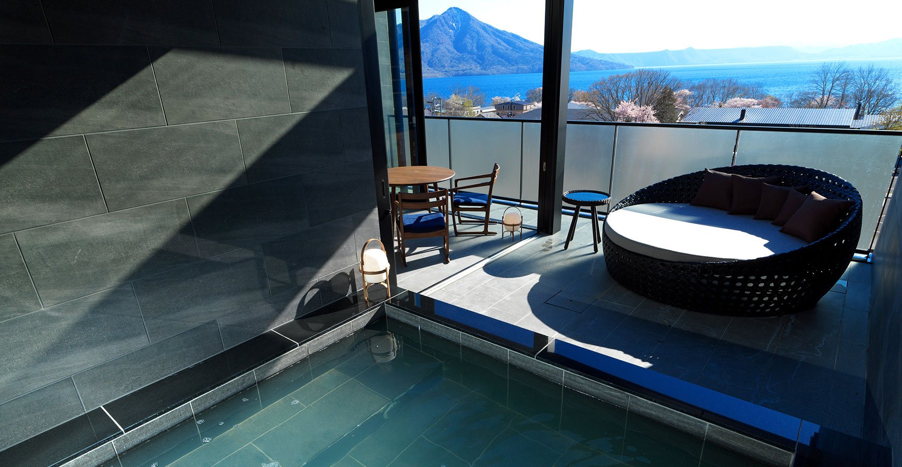 A stone open-air onsen on a suite's terrace at Ao no Za, a daybed beside it and Lake Shikotsu and a volcano beyond the rail