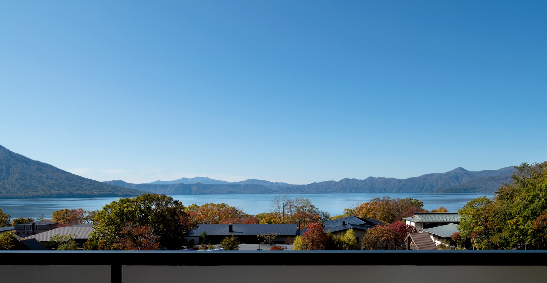 Lake Shikotsu and its far mountains seen over the ryokan's roofs and the autumn trees, under a clear sky