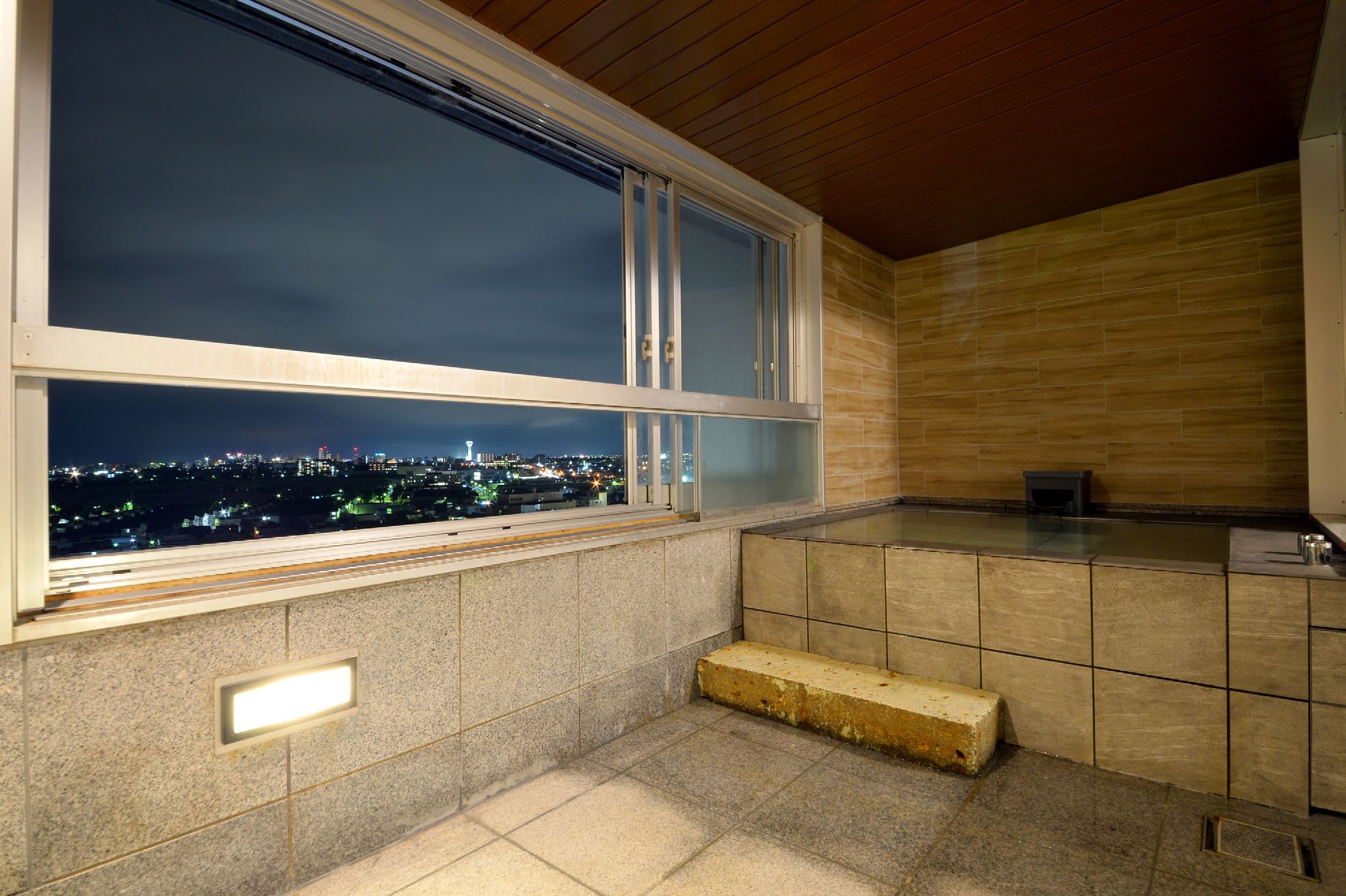 A stone bath beside a wide window onto the lights of Hakodate at night, in a guest room of Bourou Noguchi Hakodate