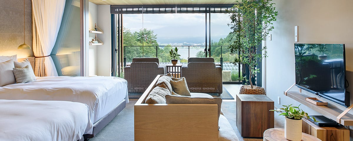 A room with a bed and a sofa, armchairs on the terrace beyond the glass and the lake below