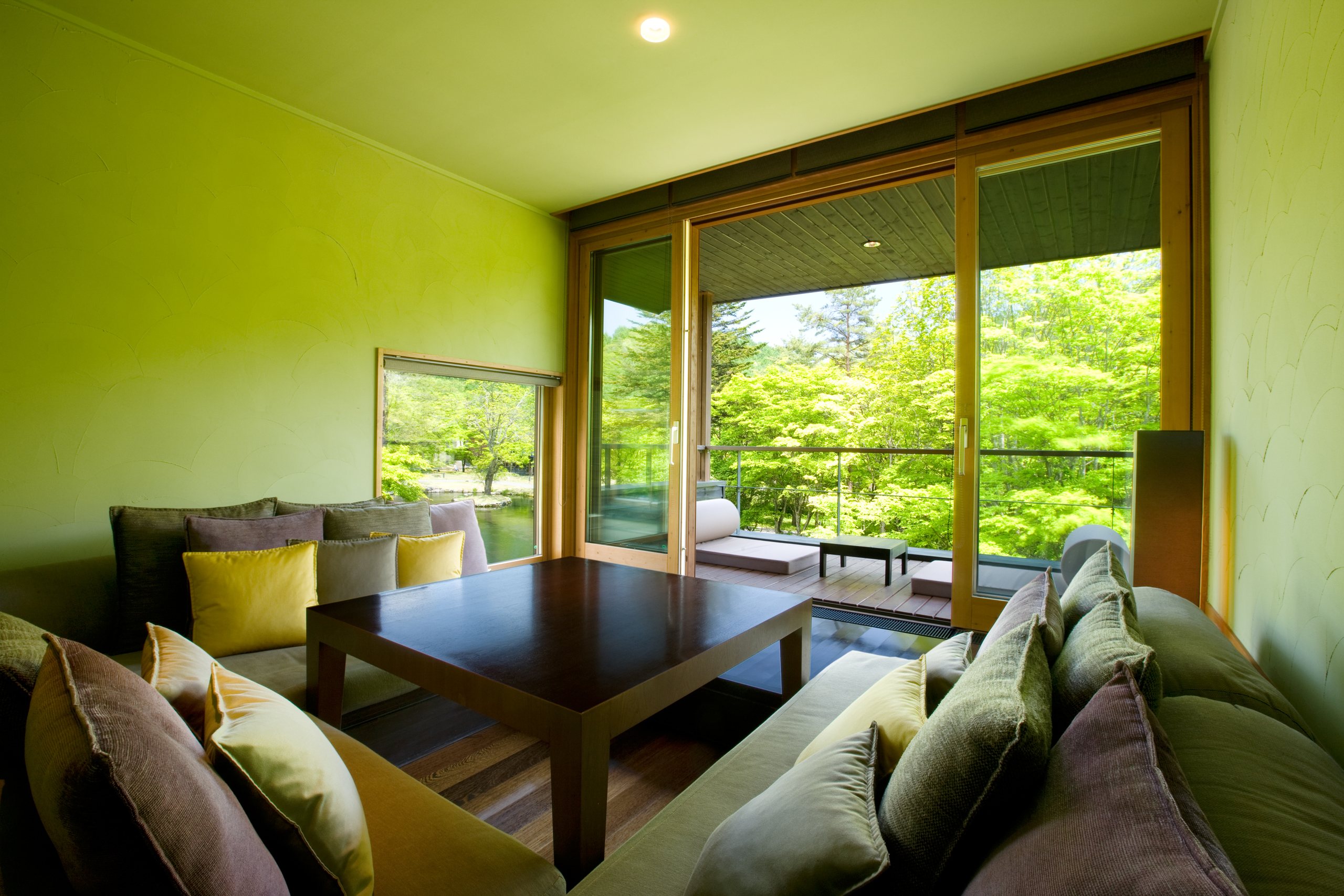 A sitting room with green walls and a low table, a wall of glass onto the trees