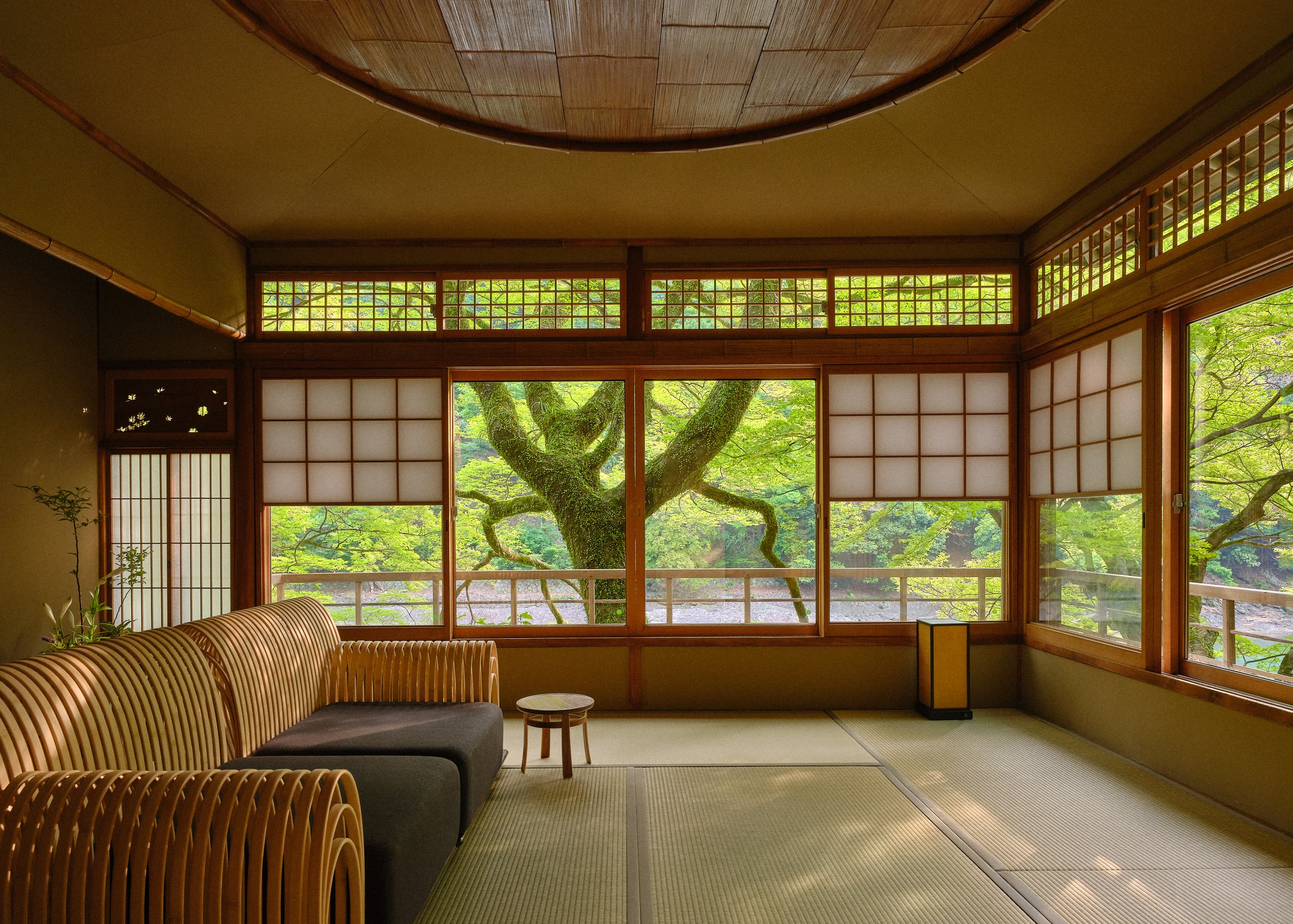 A tatami room with cane sofas beneath a round timber ceiling, its windows open onto a green tree