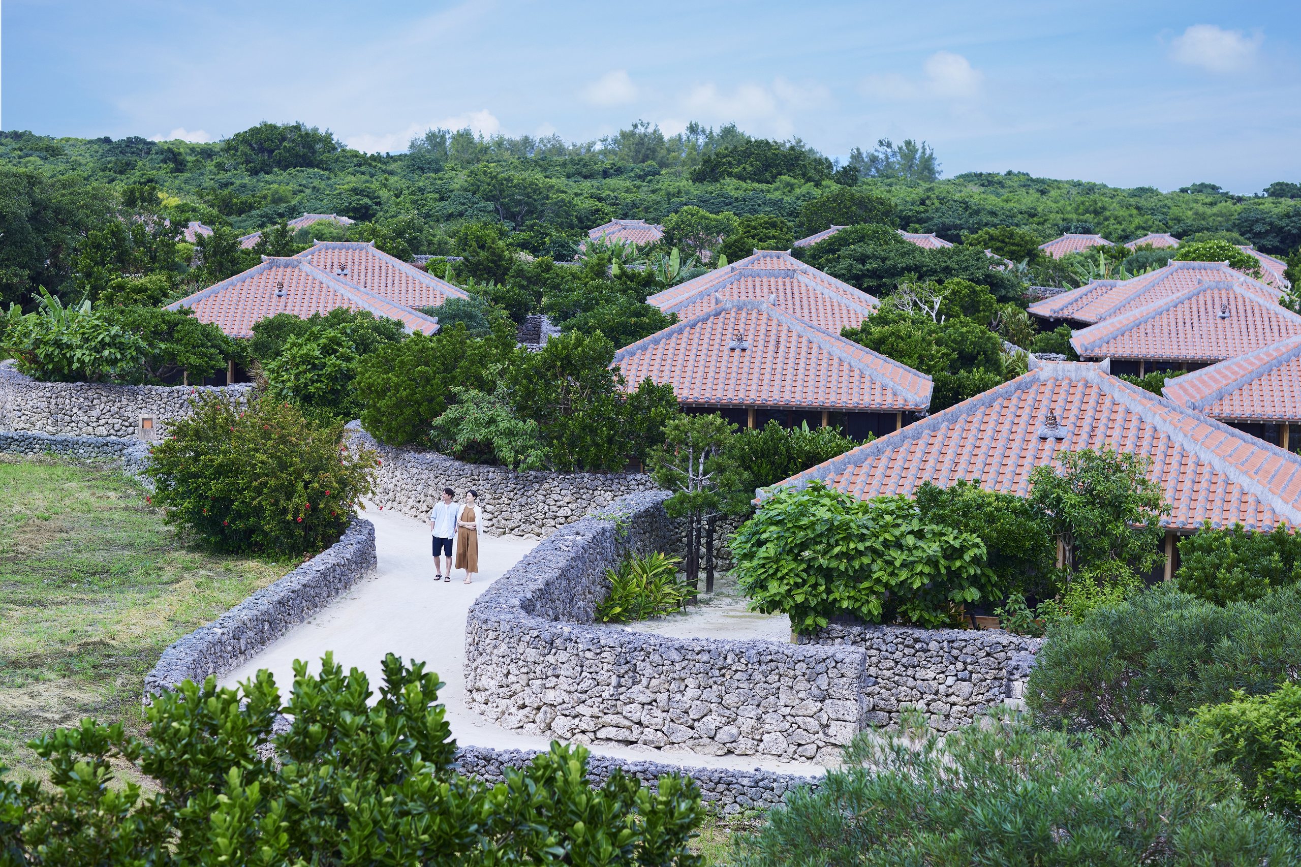 Red tiled roofs among green trees and coral stone walls, two guests on a white sand lane