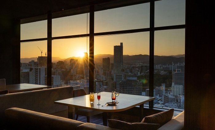 A table and chairs before tall windows over Hiroshima at sunset, in the hotel's lounge