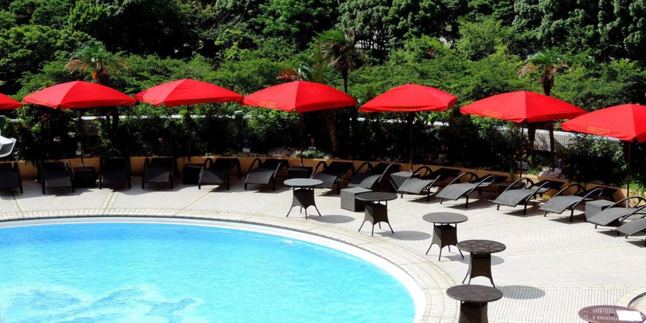 An outdoor pool with loungers under red parasols, green trees behind