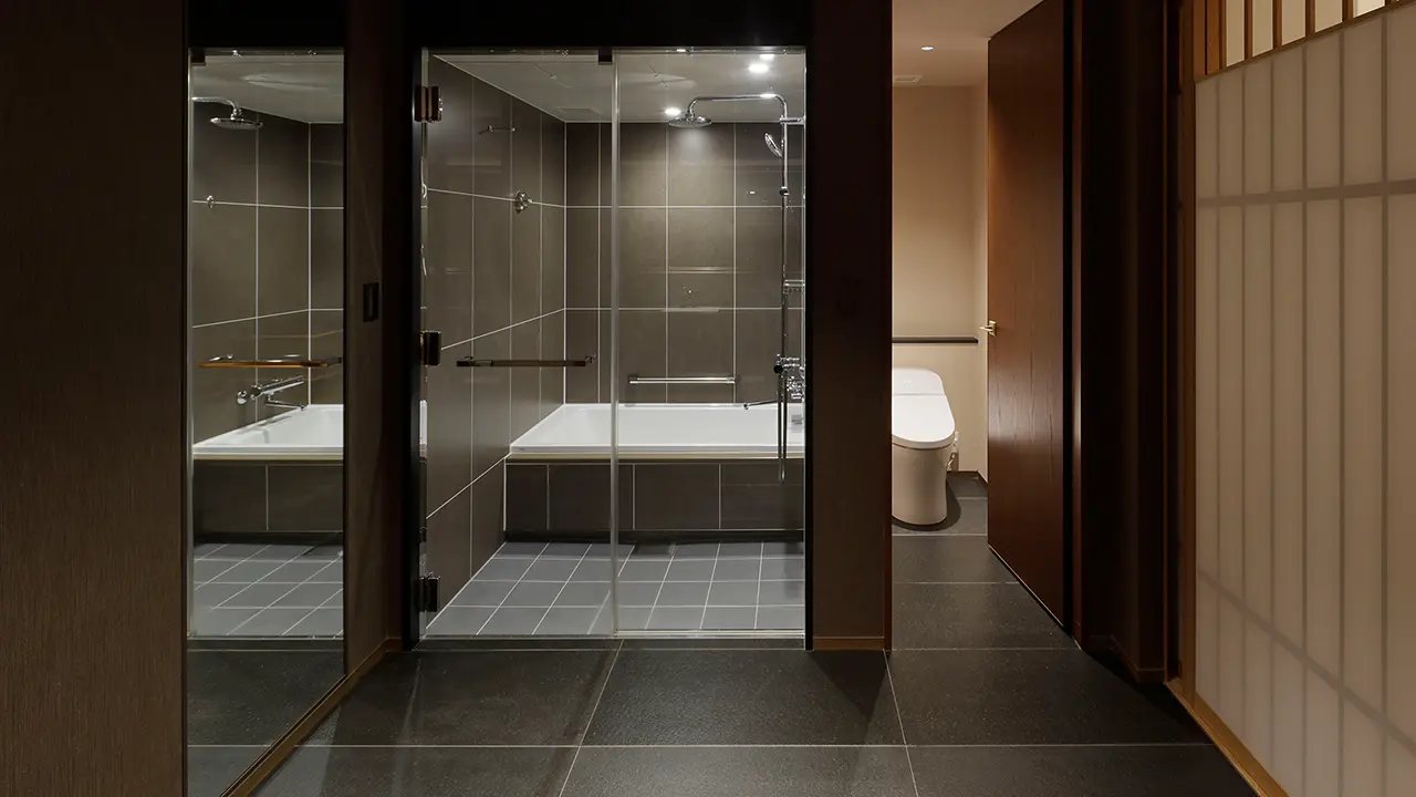 A junior suite bathroom in dark stone, a deep tub and shower behind a glass wall