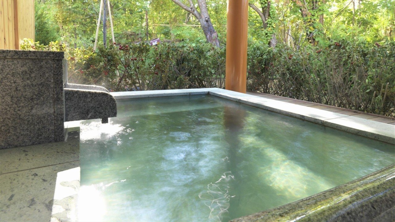 An open-air onsen pool edged in stone among green trees, at Jozankei Tsuruga Mori no Uta