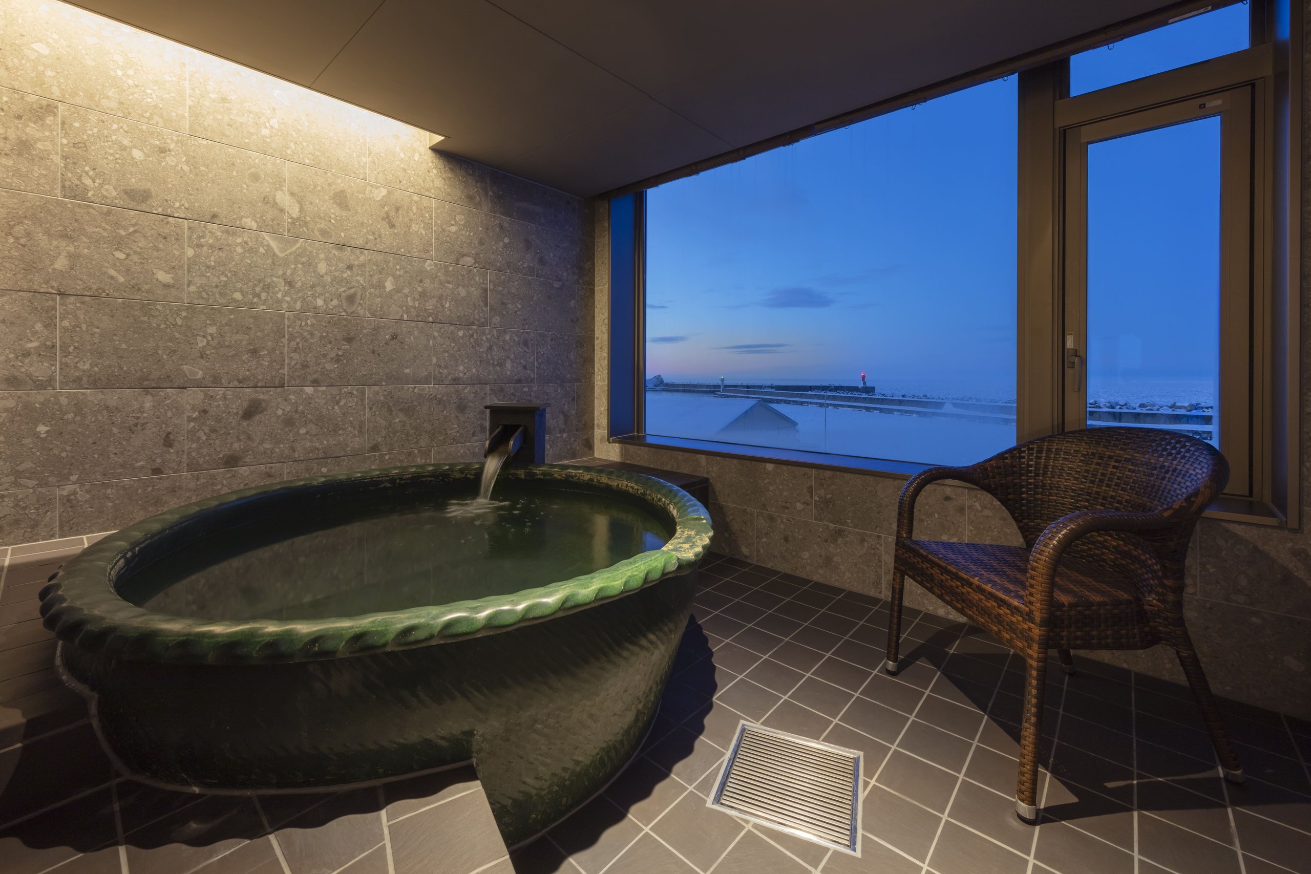 A round stone onsen and a wicker chair on a guest room terrace at Kitakobushi Shiretoko, the Okhotsk Sea beyond the glass