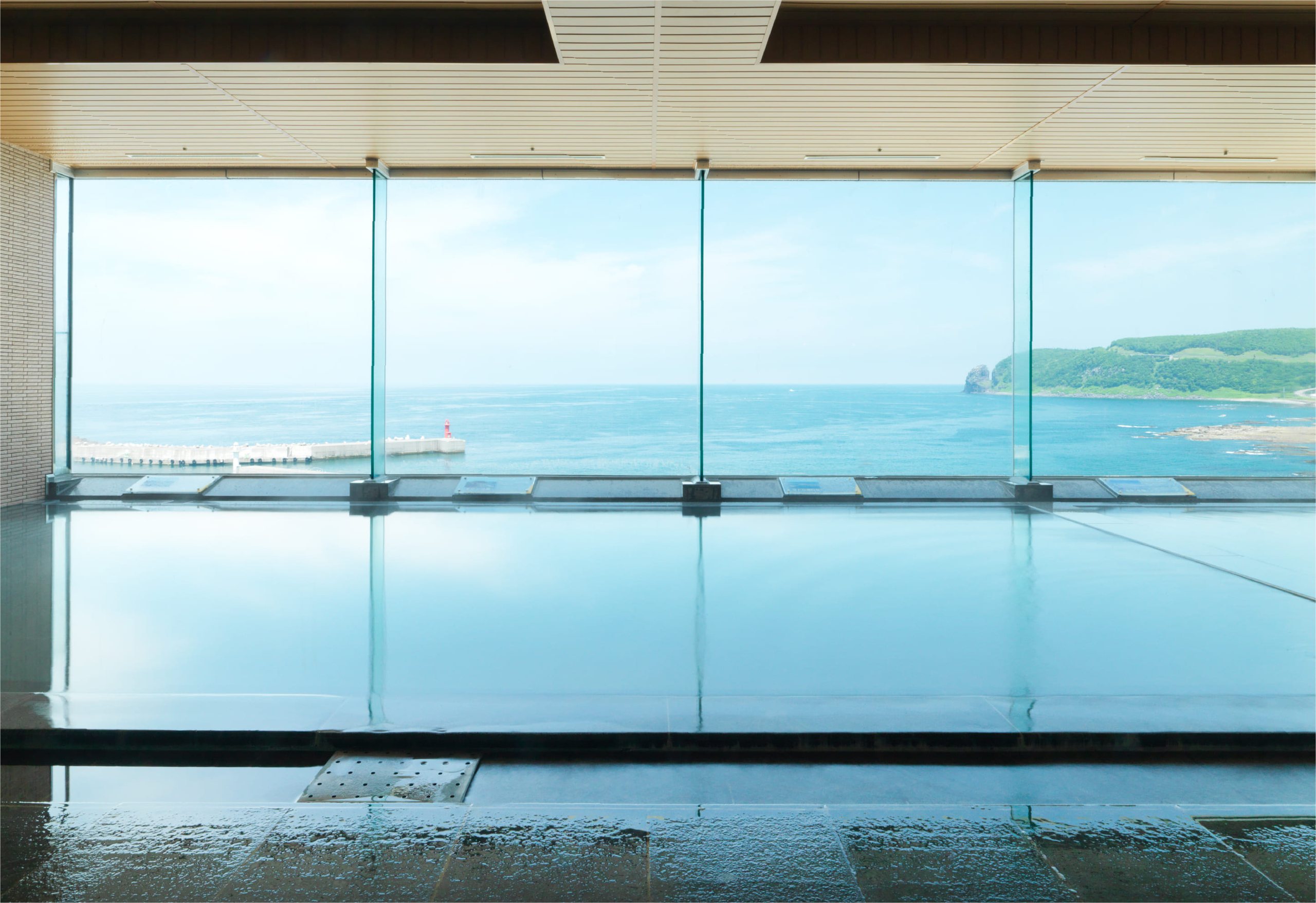 The onsen hall at Kitakobushi Shiretoko, a long pool before a glass wall onto the sea and a green headland