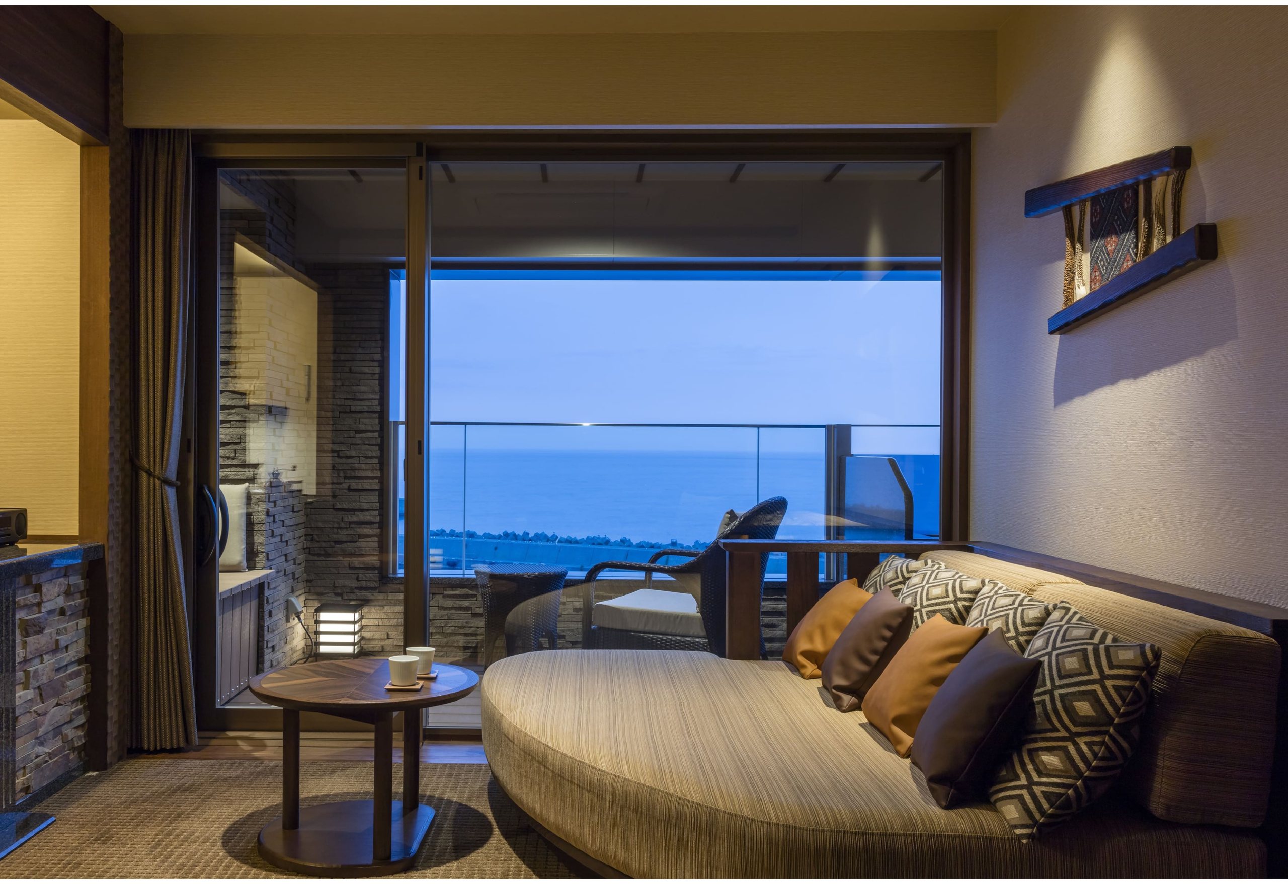 A guest room with a curved sofa and a round table before a window onto the sea at dusk