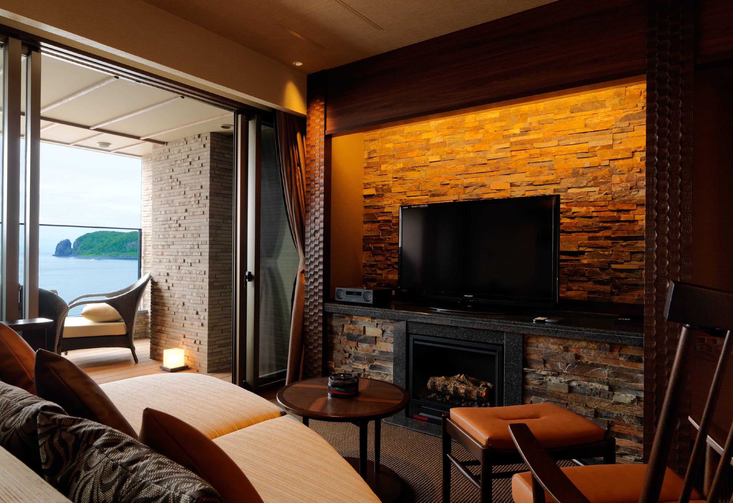 A guest room with a stone hearth and armchairs, the terrace and the sea beyond the glass