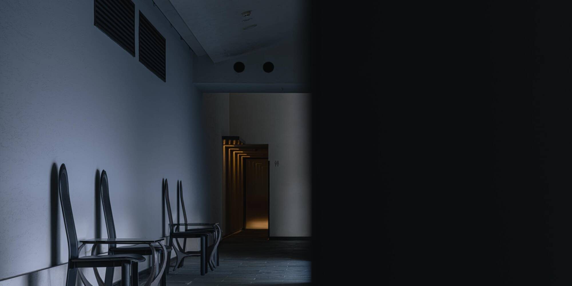 A dim corridor at Kuramure, dark chairs against a pale plaster wall and a lit doorway at the far end