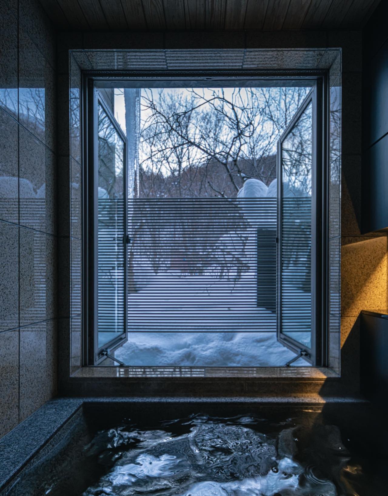 A stone onsen bath beneath a tall window onto a wooden deck and bare trees, in a room at Kuramure