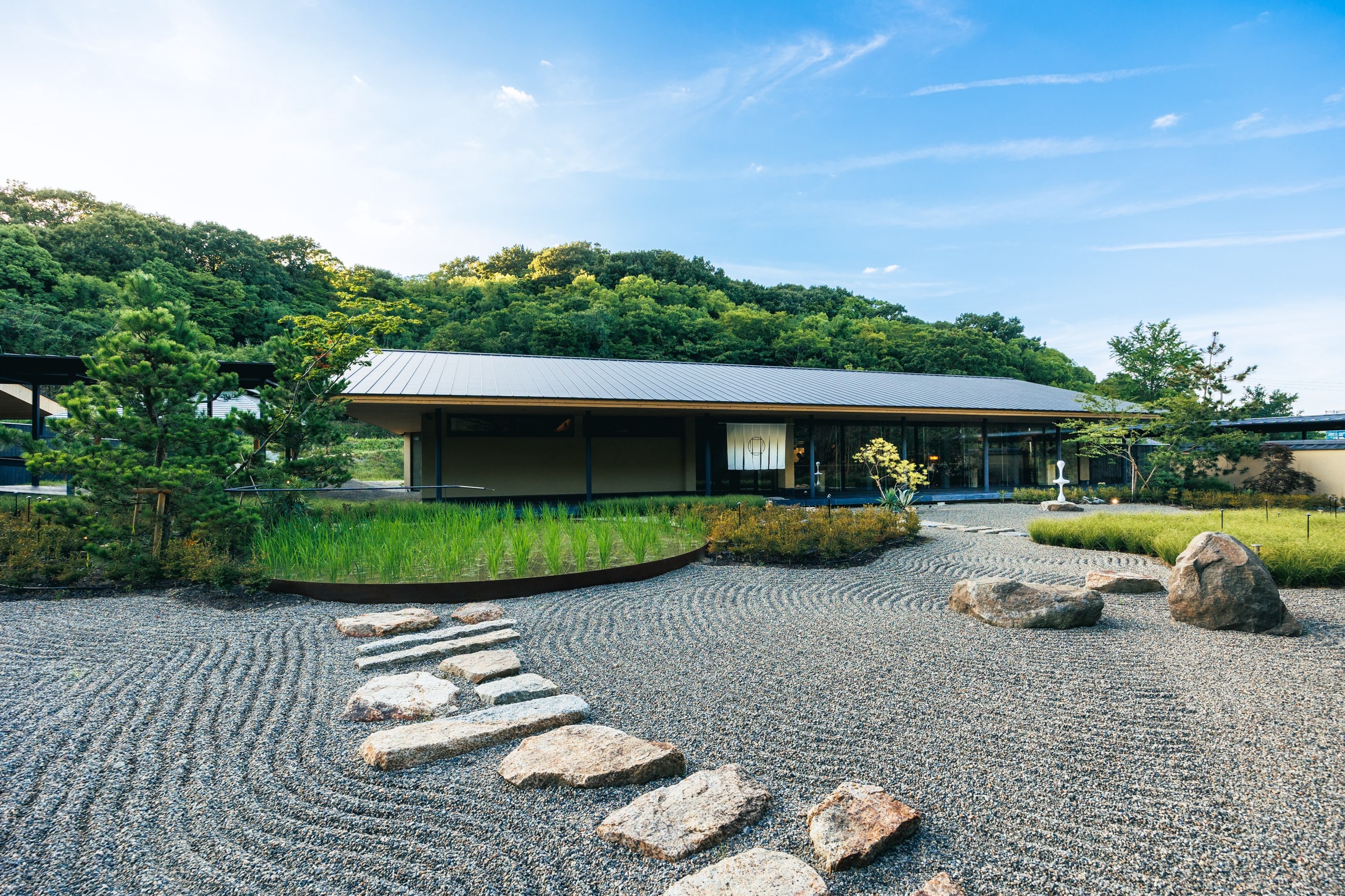 Stepping stones across raked gravel before the ryokan's long low roof, green hills behind
