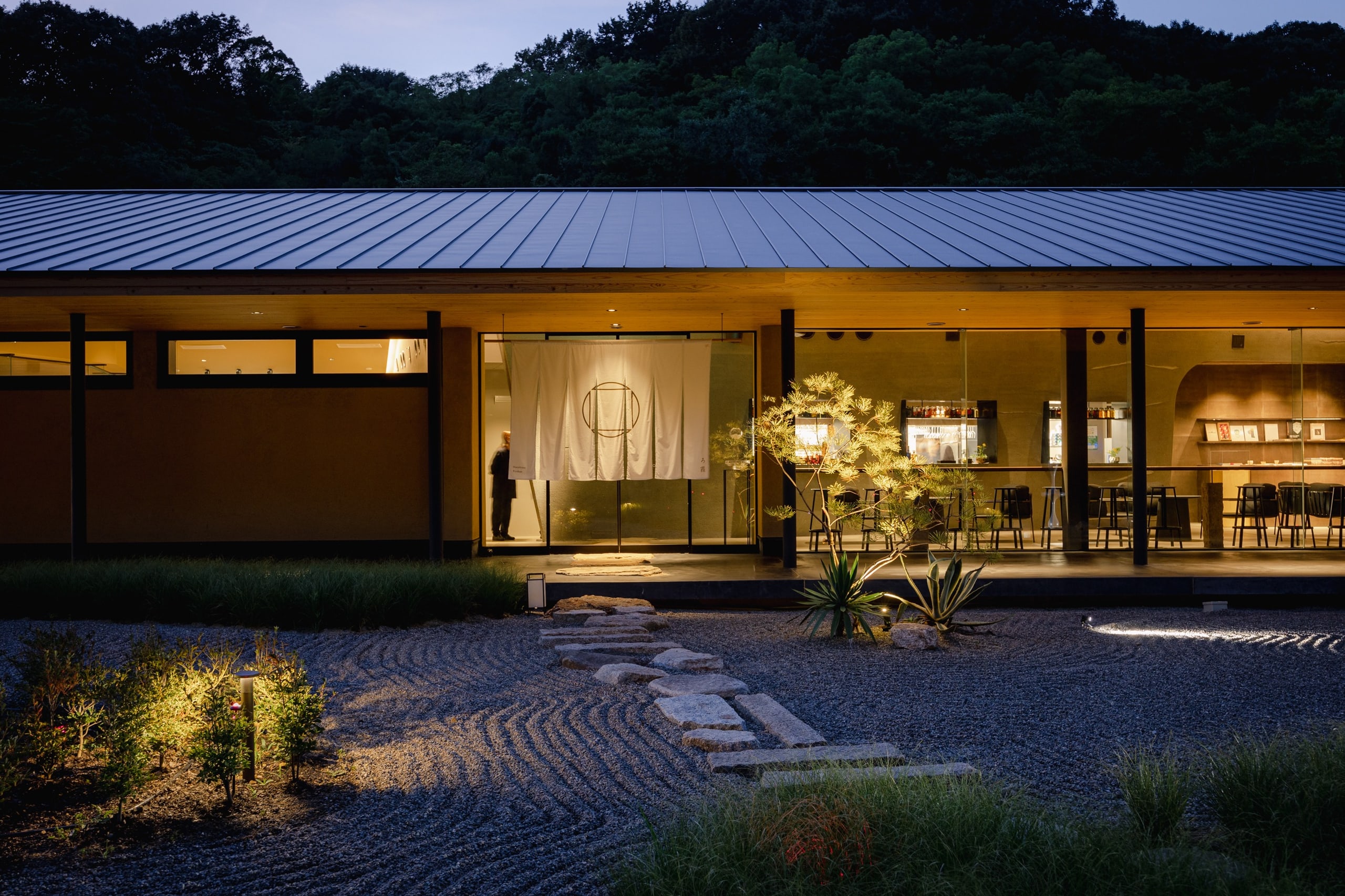 The ryokan's roof lit at night, a noren at the door and raked gravel before it