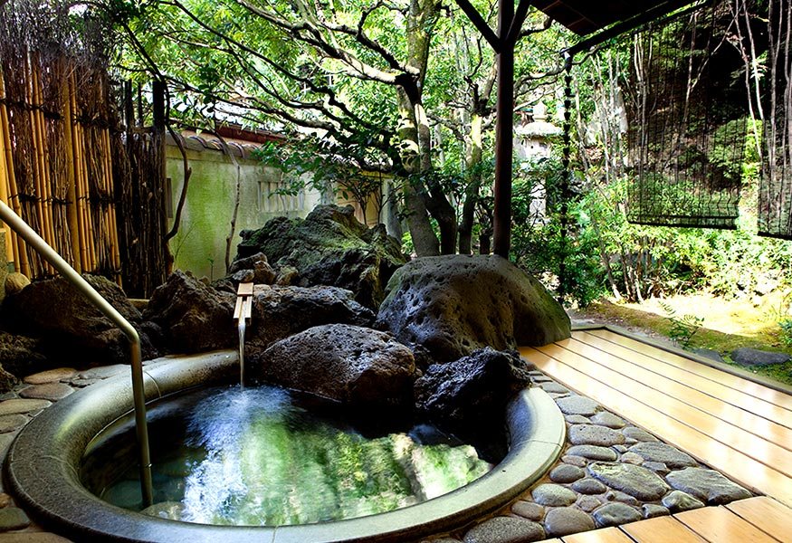 A round onsen set in rock under the trees of a garden, bamboo and a wooden deck beside it