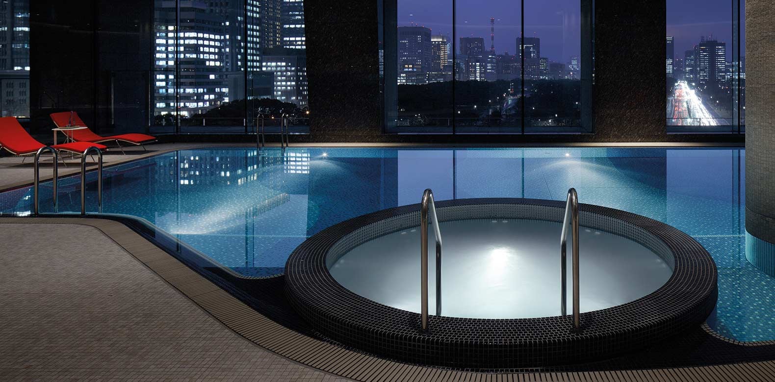An indoor pool and a round whirlpool at night, the lit towers of the city in the windows beyond