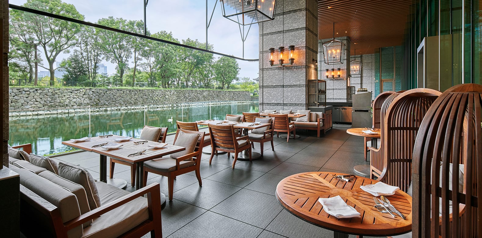 Tables and armchairs on a covered terrace beside the moat and its stone walls, trees beyond