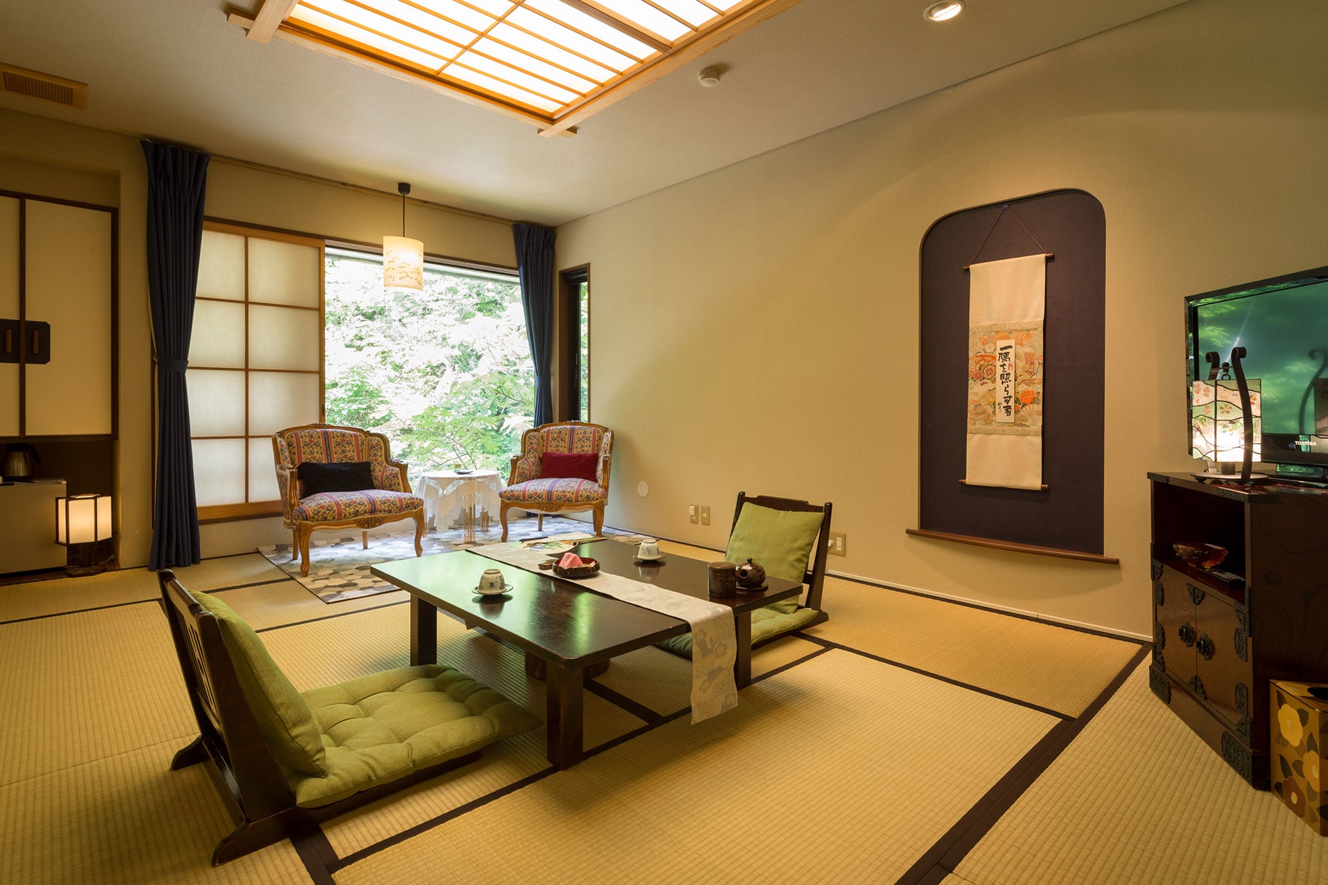 A tatami room with a table and chairs, a hanging scroll in an arched alcove and a window onto the trees