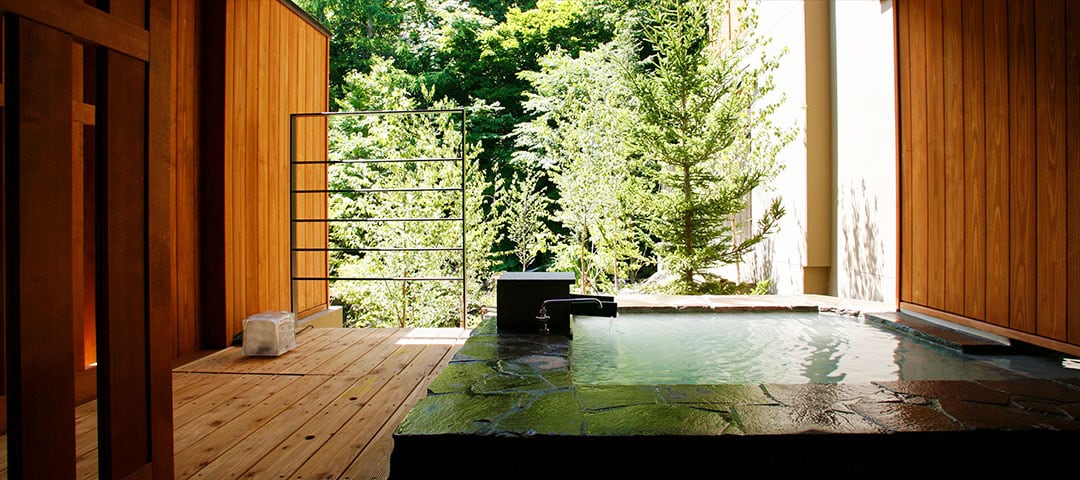 A room's own open-air bath in stone on a timber deck, green trees beyond
