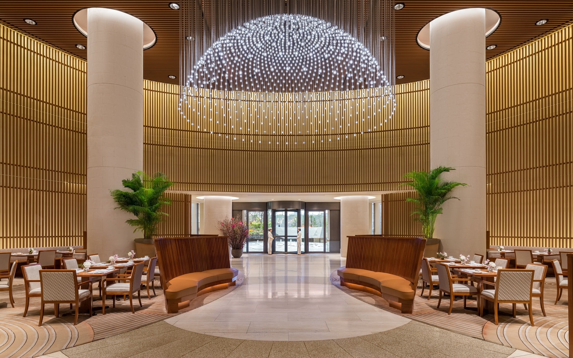 A round lobby of gold timber screens beneath a chandelier of beaded lights, tables laid for tea