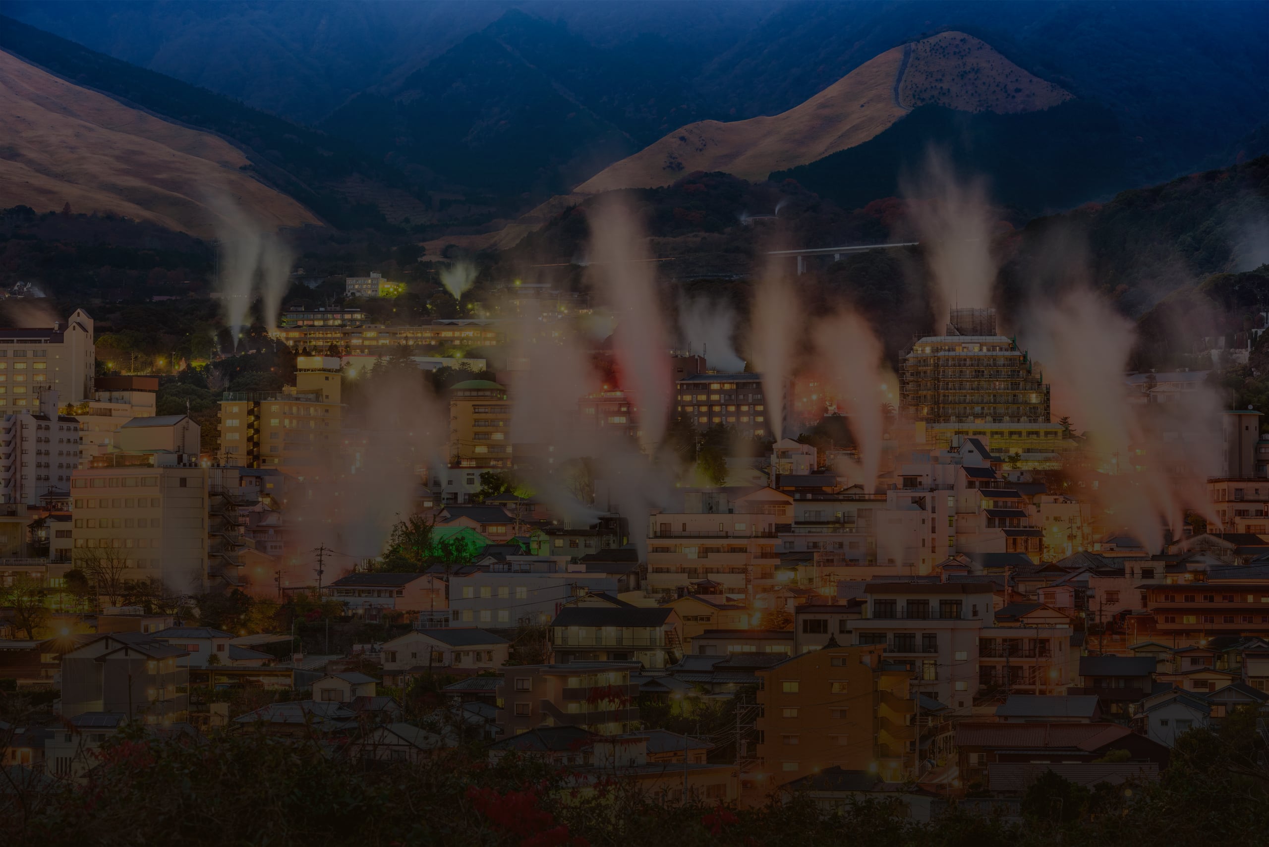 Columns of steam rising over the lit hillside town of Beppu at dusk, mountains behind