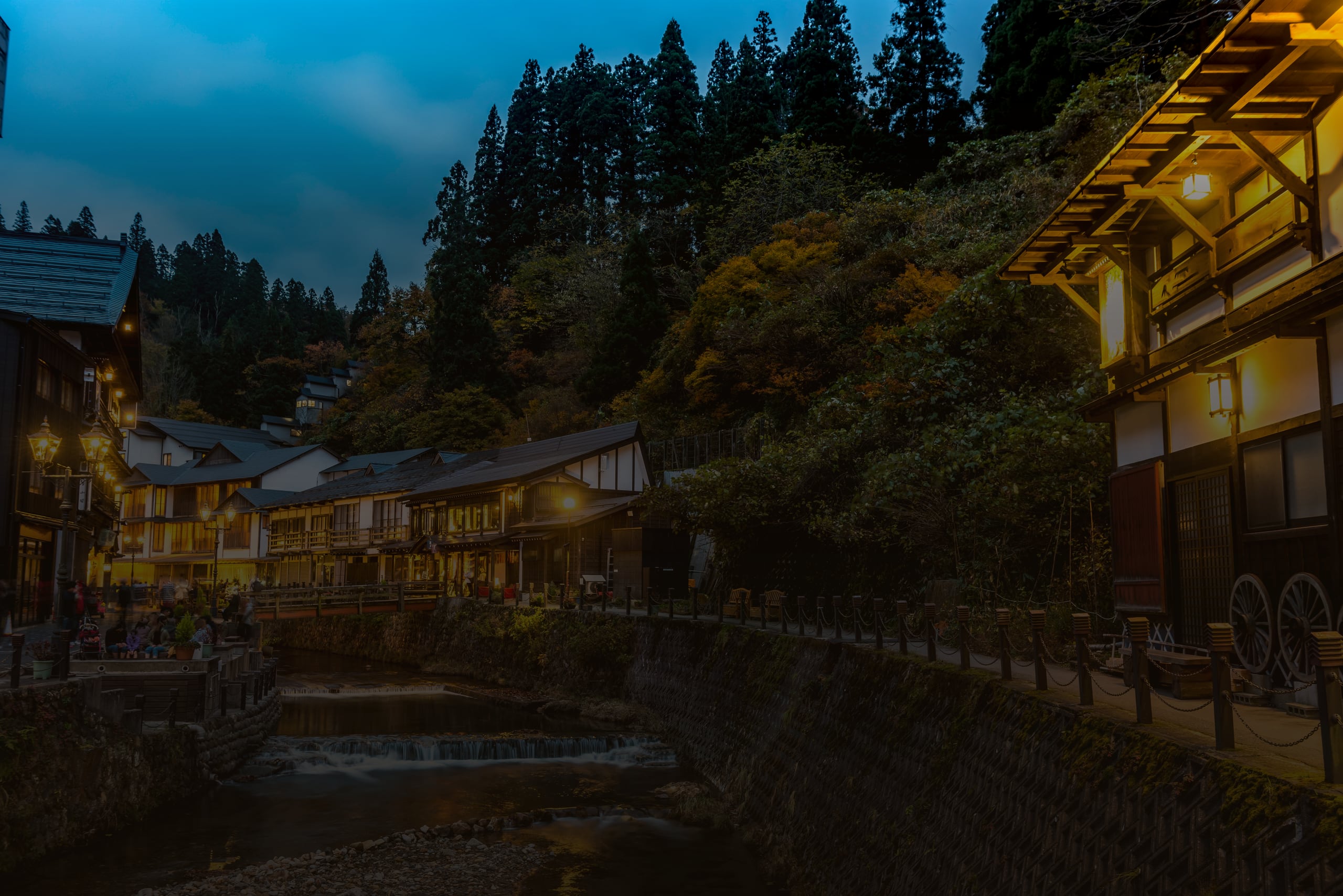 The wooden inns of Ginzan Onsen lit along the river at dusk, forest on the slope above