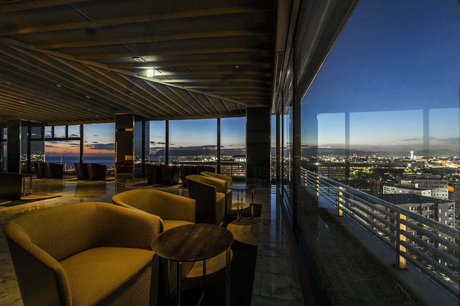 A lounge of low armchairs behind a wall of glass, the lights of Hakodate below at dusk