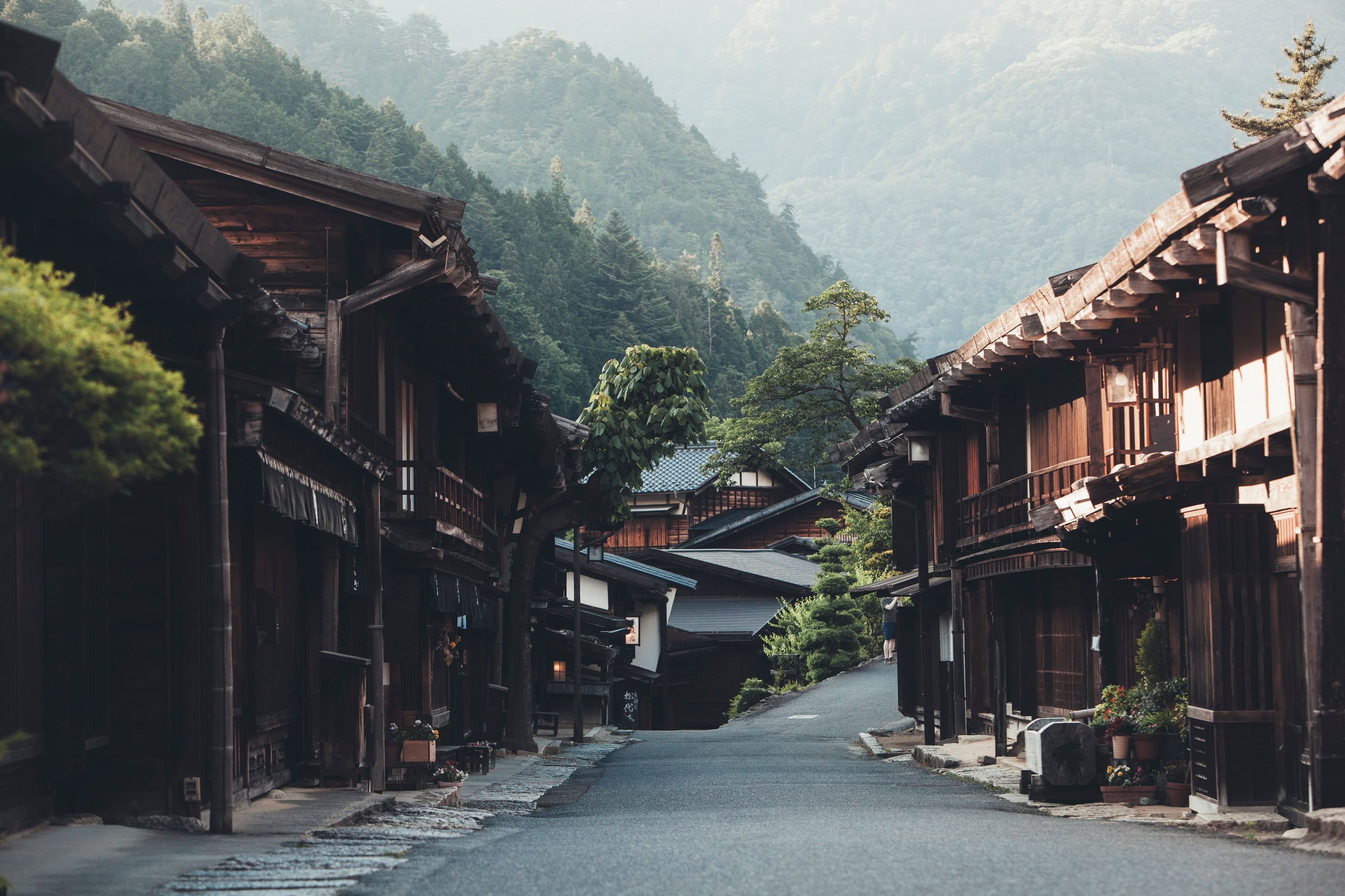An empty paved street through a Nakasendo post town in the Kiso Valley, wooden houses along it and misty green hills behind