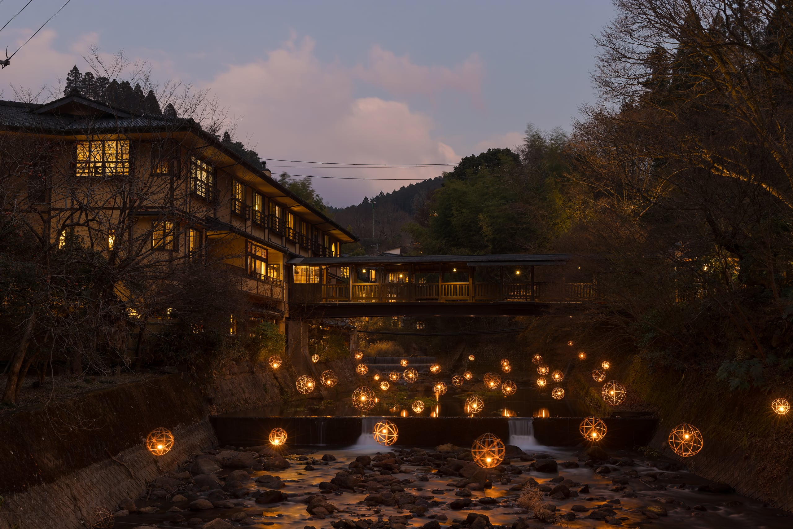 Timber ryokan buildings and a covered wooden footbridge over a river lined with lanterns at dusk