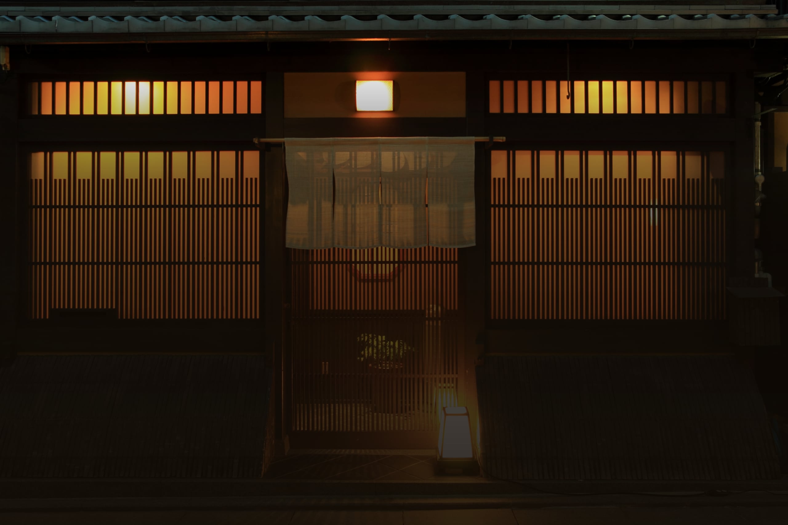 The latticed wooden front of a Kyoto townhouse at night, a noren curtain at the door and a lantern lit on the step