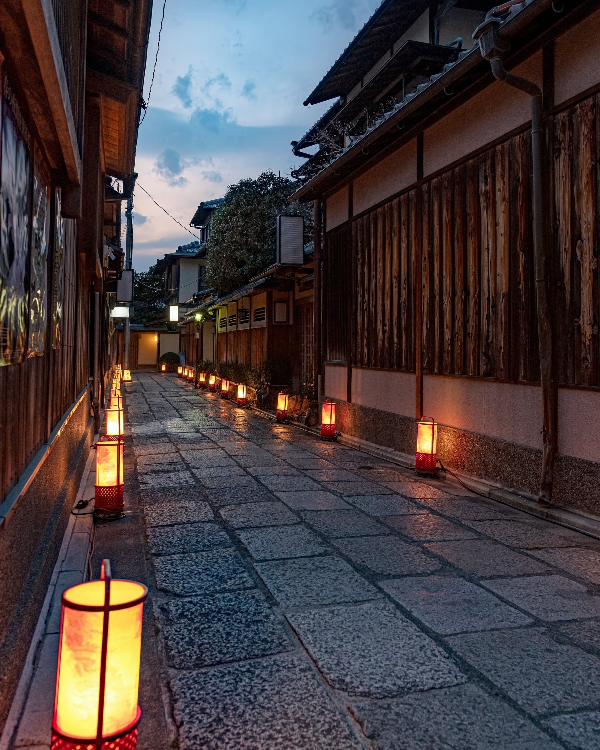 A narrow stone-flagged lane between wooden walls in Higashiyama, Kyoto, lit by paper lanterns set along the ground at dusk
