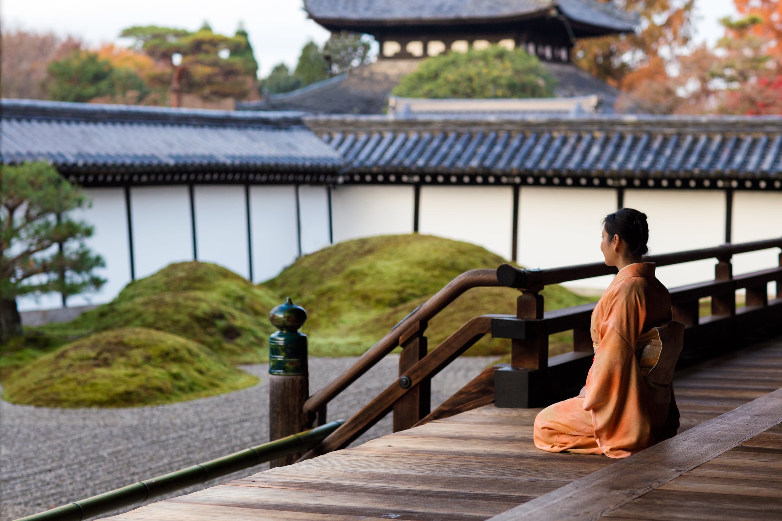 A woman in an apricot kimono kneeling on a temple veranda in Kyoto, looking out over mounds of moss and raked gravel below a long white wall