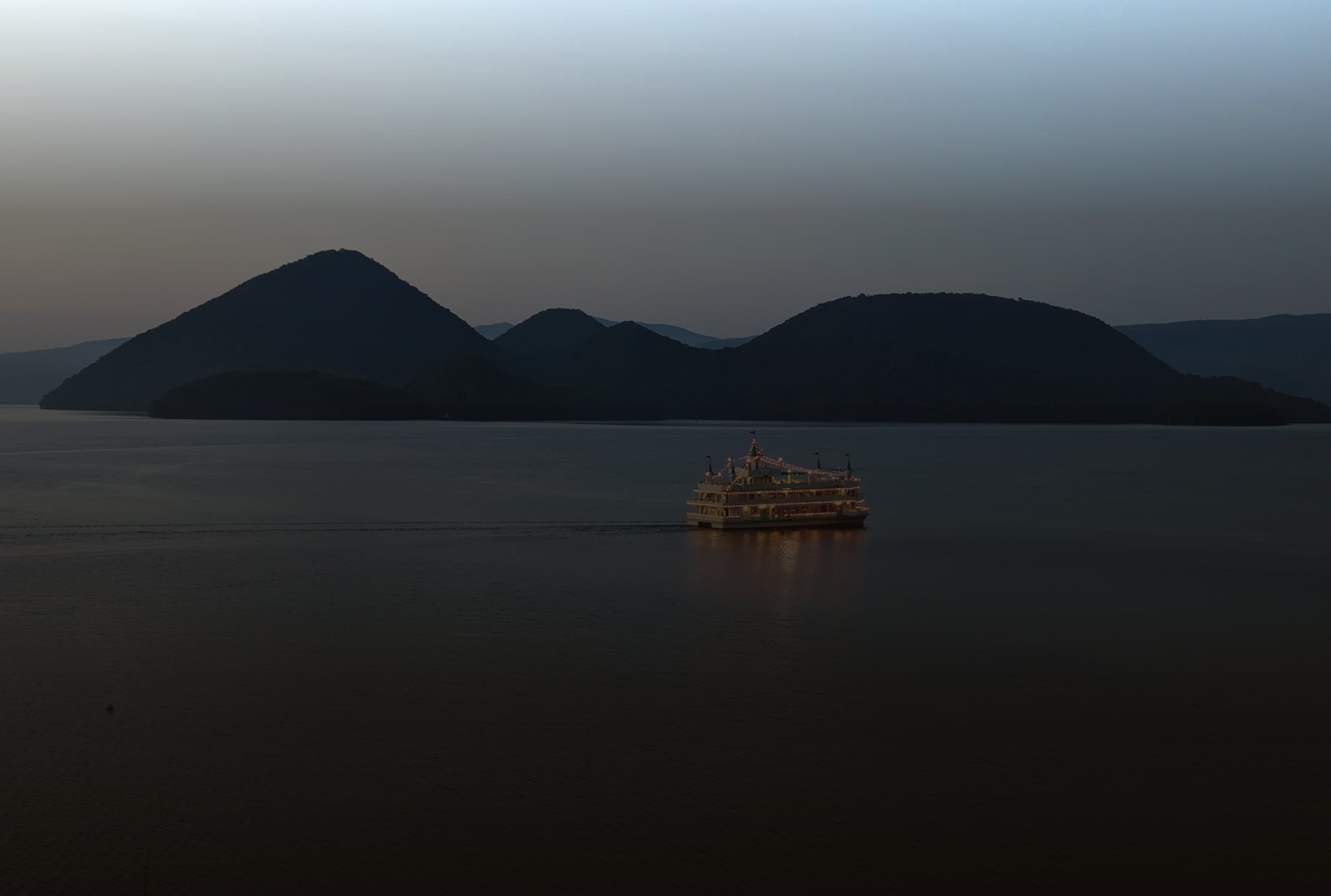 A lit sightseeing boat crossing the calm water of Lake Toya at twilight, the Nakajima islands dark behind it