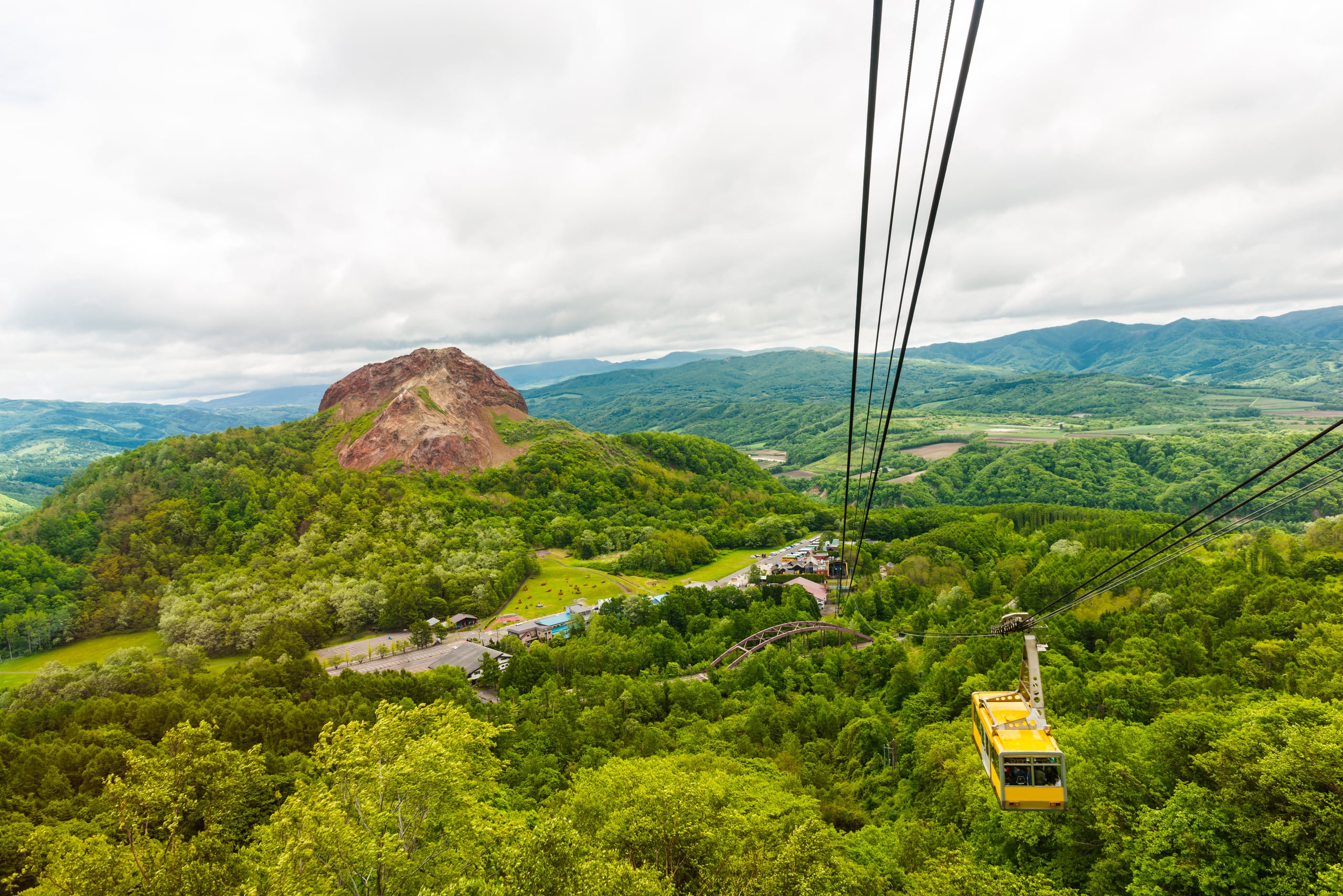 The bare red dome of Showa-Shinzan among green forest, a ropeway car crossing the slope
