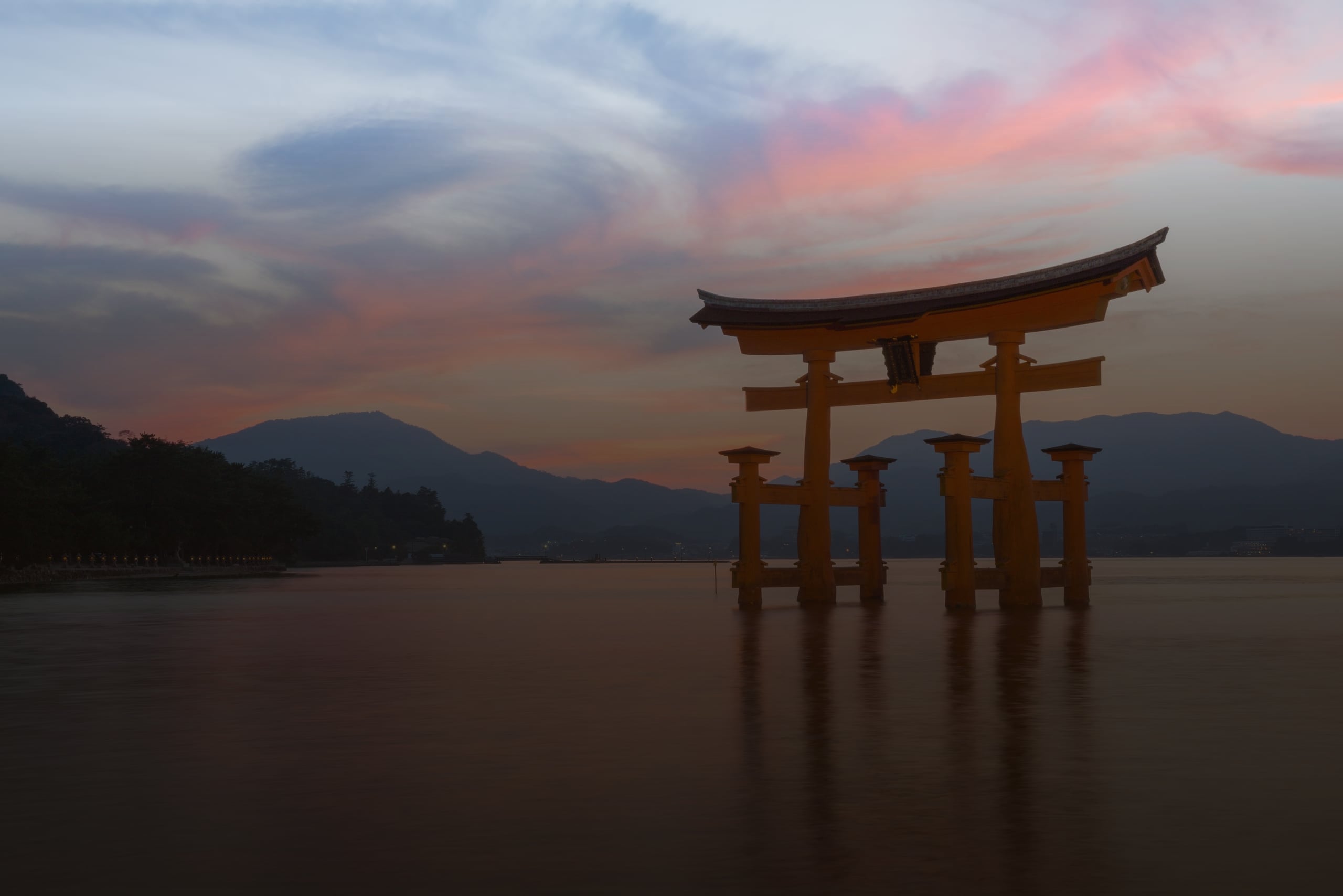 The vermilion great torii of Itsukushima Shrine standing in calm water at dusk, mountains behind